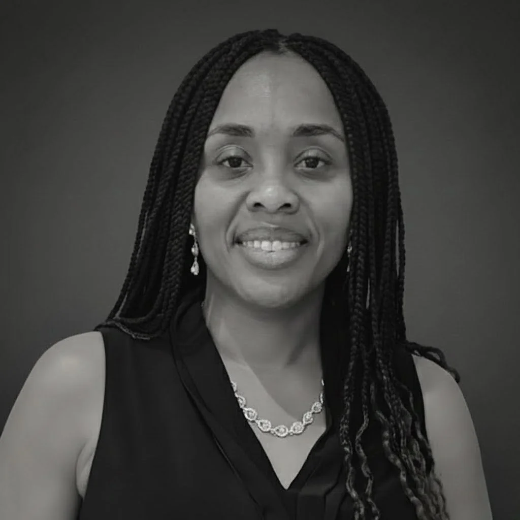 Black and white portrait of a woman with braided hair, wearing a sleeveless top, jewelry including earrings and a necklace, and smiling slightly against a plain background.