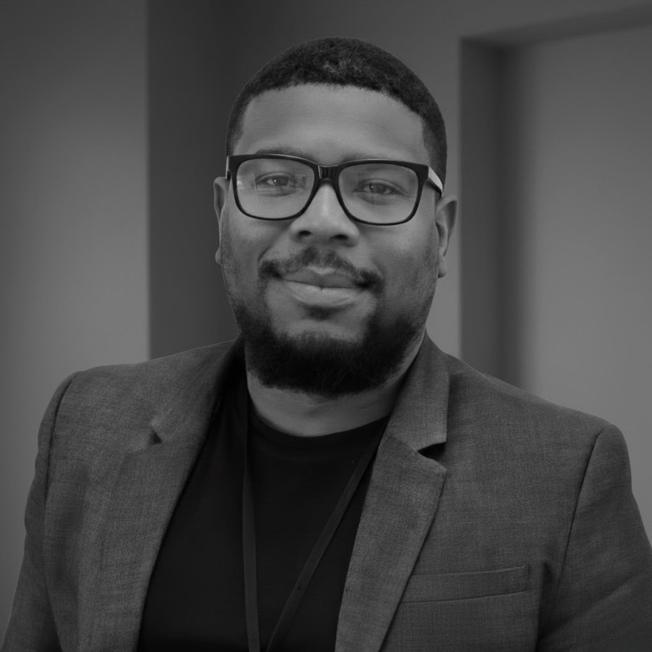 A Black man with glasses, a beard, and a mustache, wearing a blazer, smiling at the camera in a professional setting.