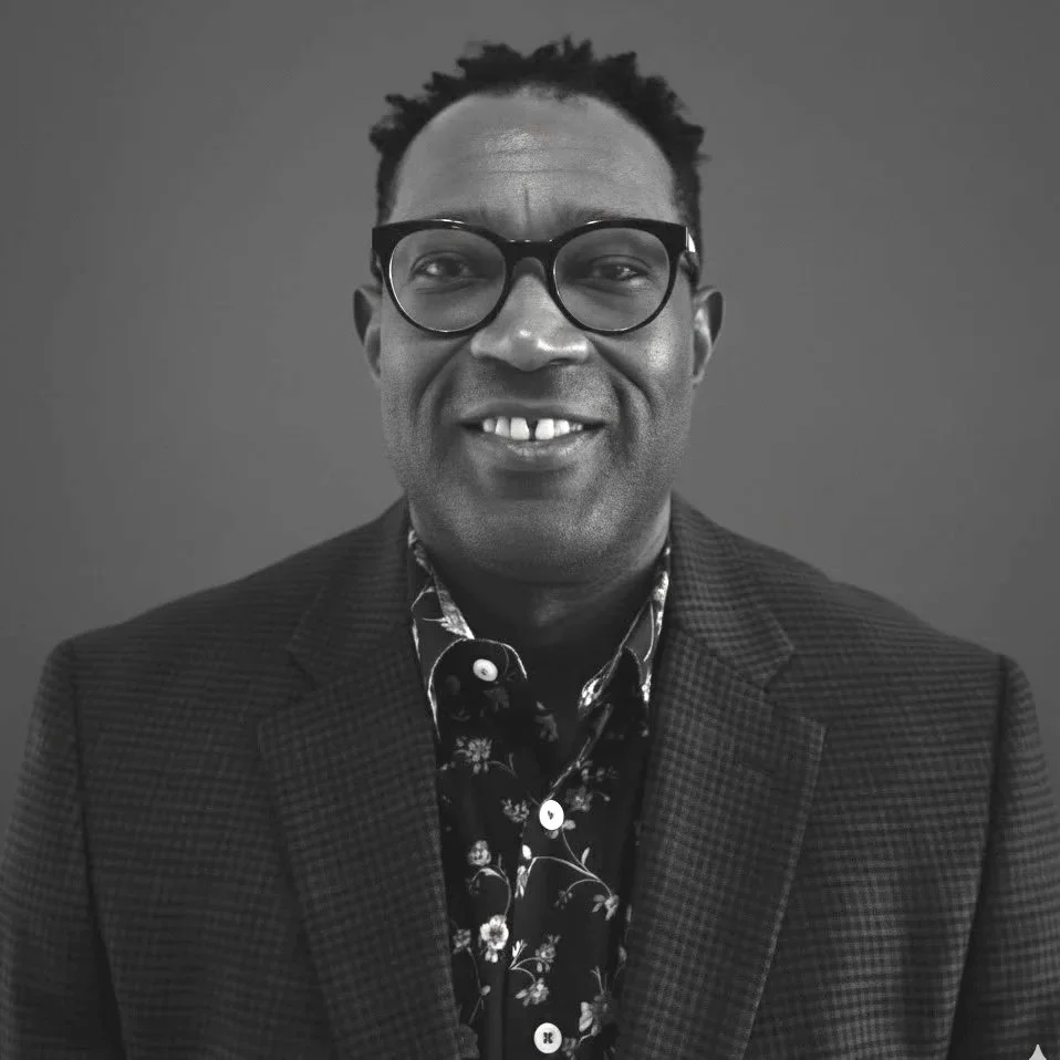 Black-and-white photo of a smiling man wearing glasses, a floral shirt, and a blazer.