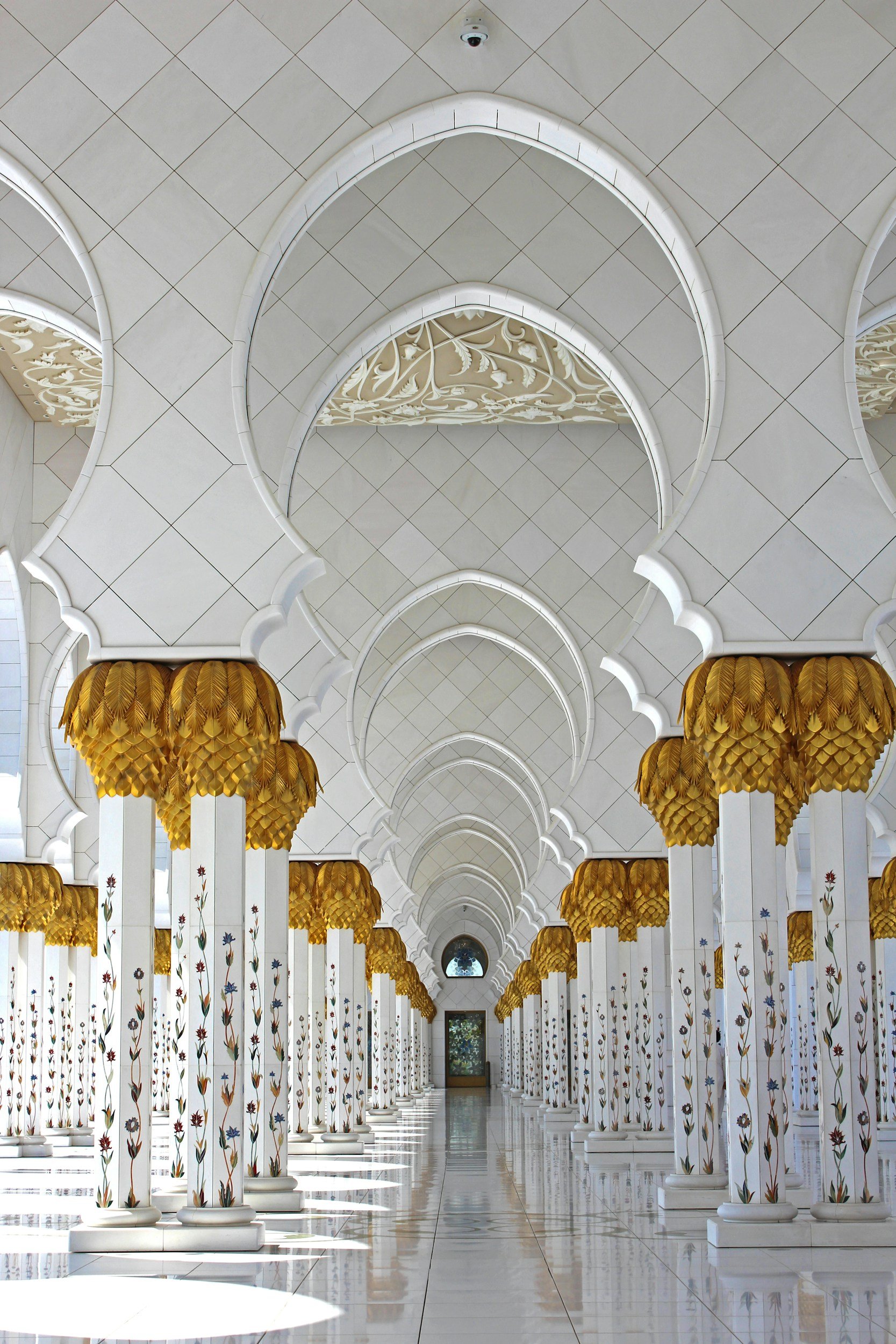 A long hallway with white columns decorated with floral patterns and golden capital tops, arched ceiling with intricate carvings, and a reflective tiled floor leading to an arched window.