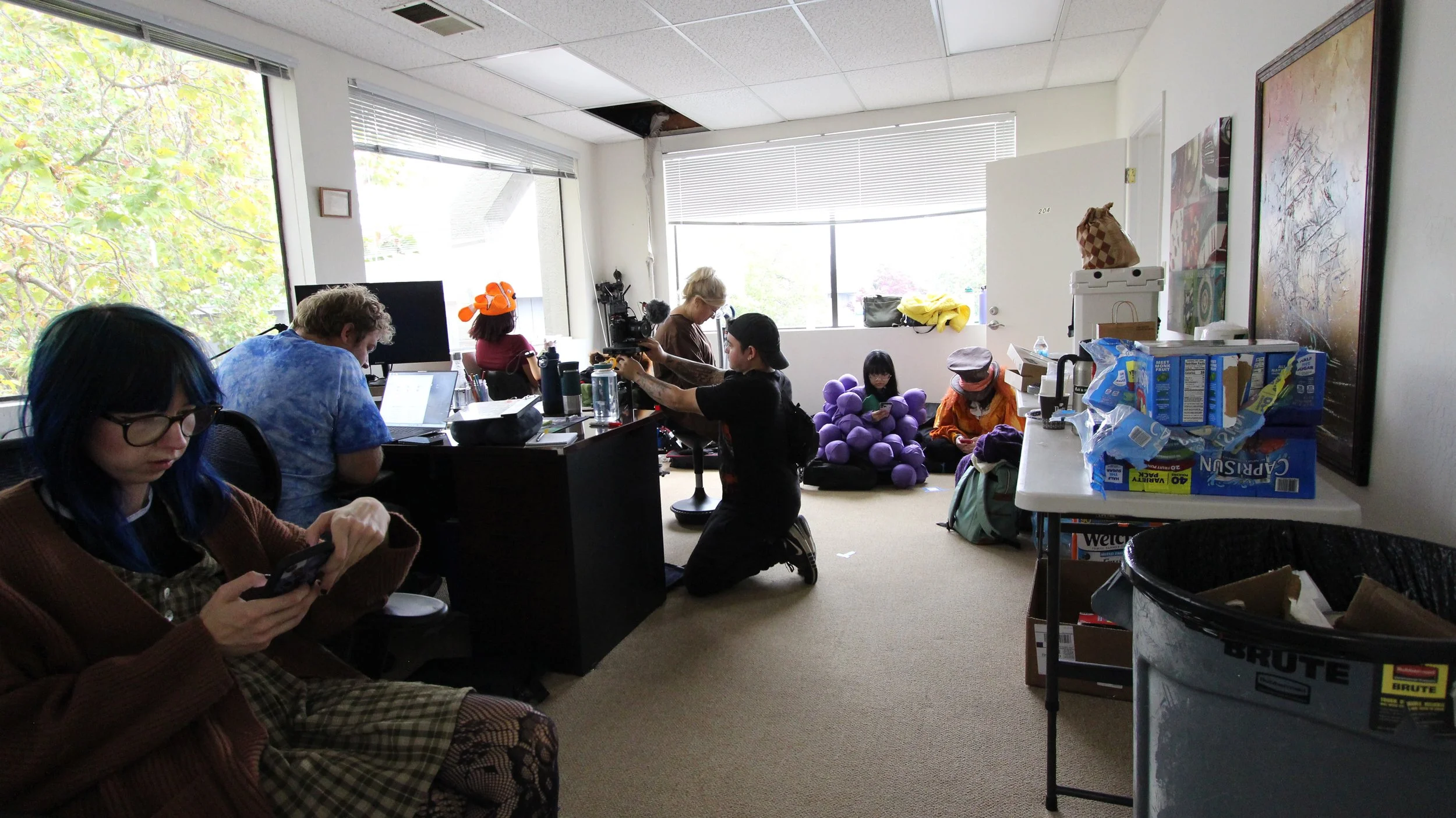 Group of people in a cluttered office, some sitting and some kneeling, engaged in various activities such as using phones, working on laptops, filming, and reading, with windowed walls showing trees outside.