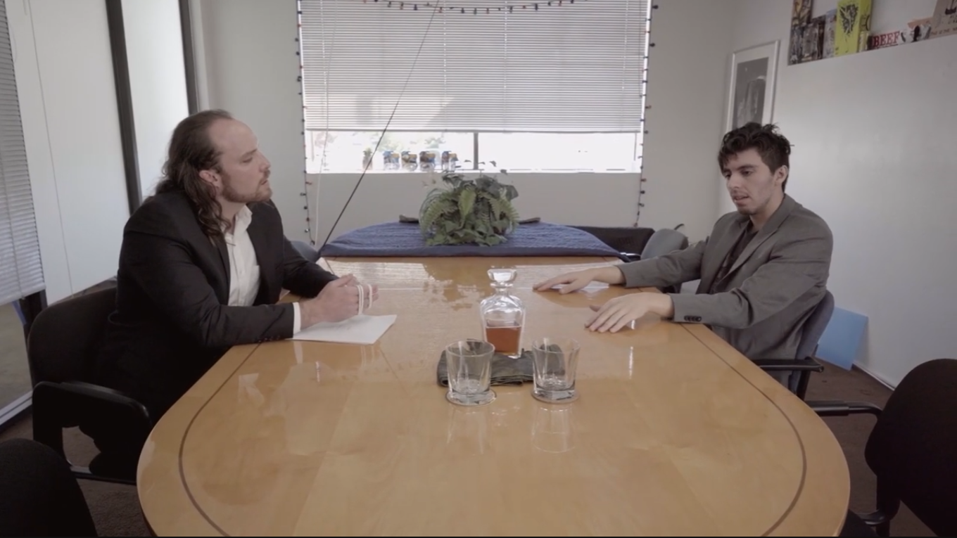 Two men sitting at a conference table in an office, having a serious conversation. One man on the left with long hair and a suit, holding a notepad. The other man on the right with short hair and a gray blazer. A glass decanter and two glasses are on the table between them. The office has a whiteboard, window with blinds, and posters on the wall.
