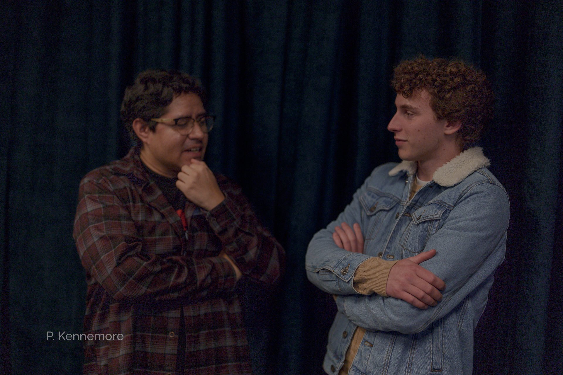 Two men face each other in front of a dark curtain, one with glasses and a plaid jacket appears to be speaking or explaining something, while the other with crossed arms listens attentively.