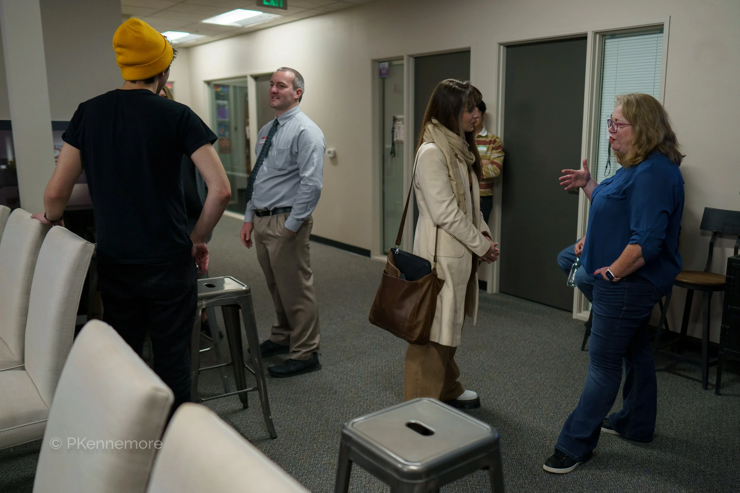 Five people having a conversation in an office or waiting area, with chairs, small tables, and a water bottle visible.