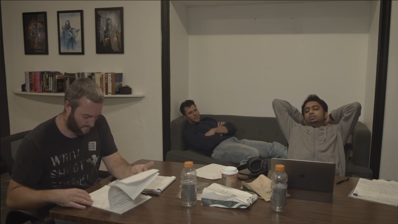 Three men in a room, one sitting at a table reading papers, one lying on a couch with arms behind head, and the third sitting on a couch with eyes closed. The table has water bottles, a laptop, and other items. The wall has framed pictures and a bookshelf.