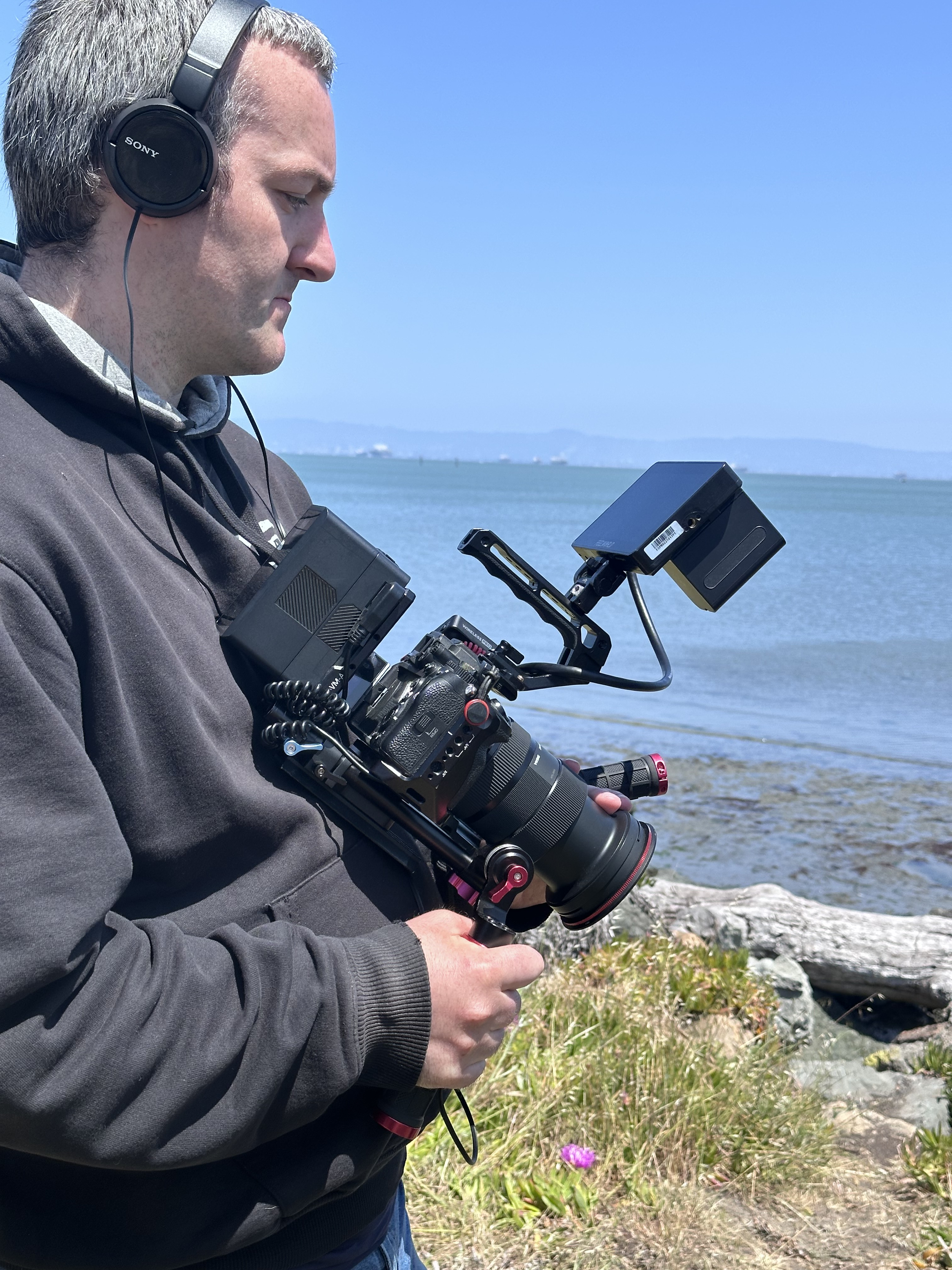 A person in a gray hoodie with headphones filming with a professional camera rig near a body of water, with rocks, grass, and a log in the background under a clear blue sky.