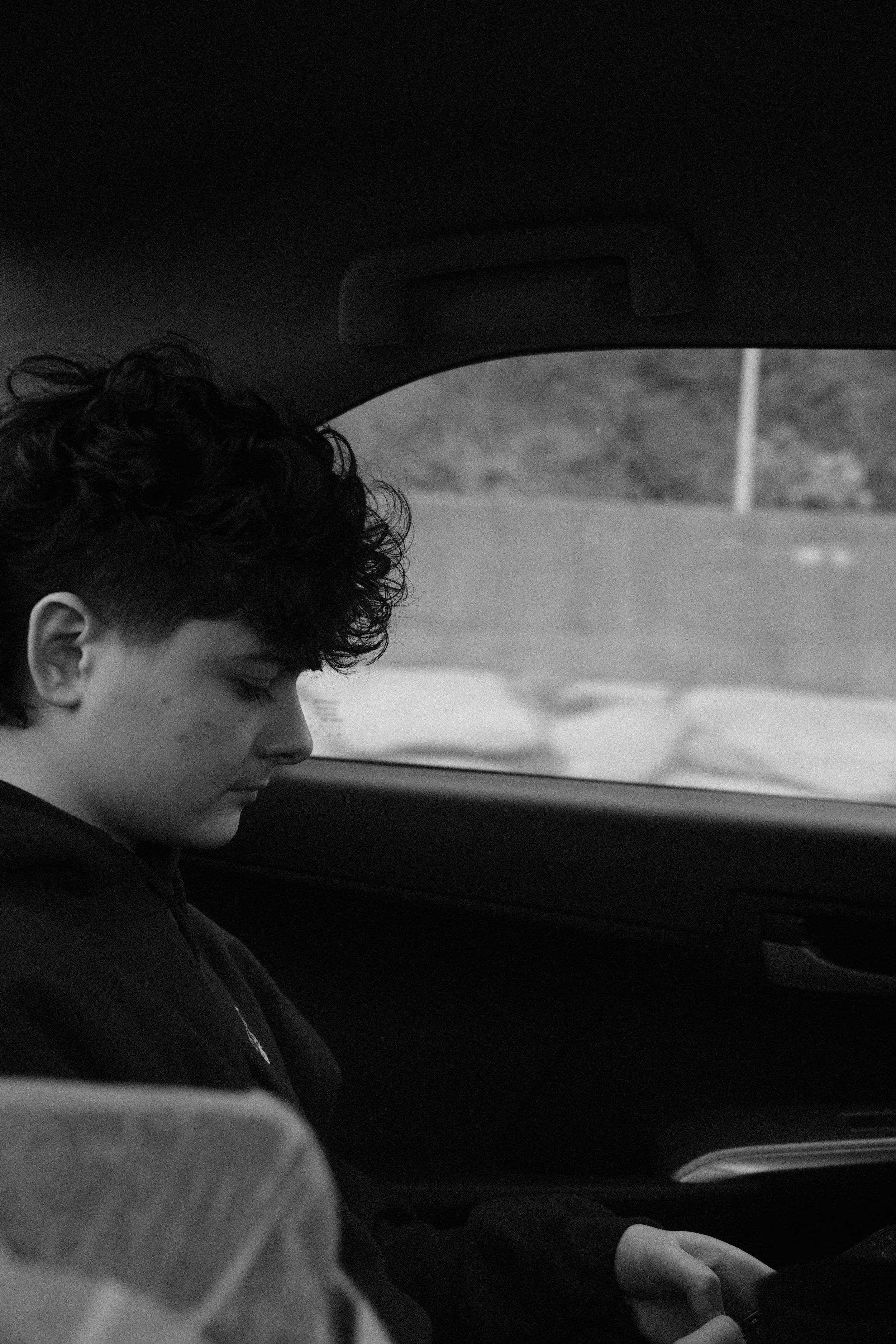 A young person with dark curly hair sitting inside a car, looking at a mobile device, with a blurred outdoor scene outside the window.