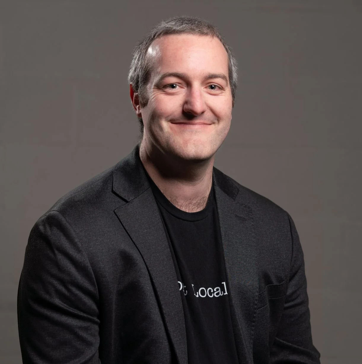 Headshot of a smiling man with short hair, dressed in a dark blazer and t-shirt, against a neutral background.
