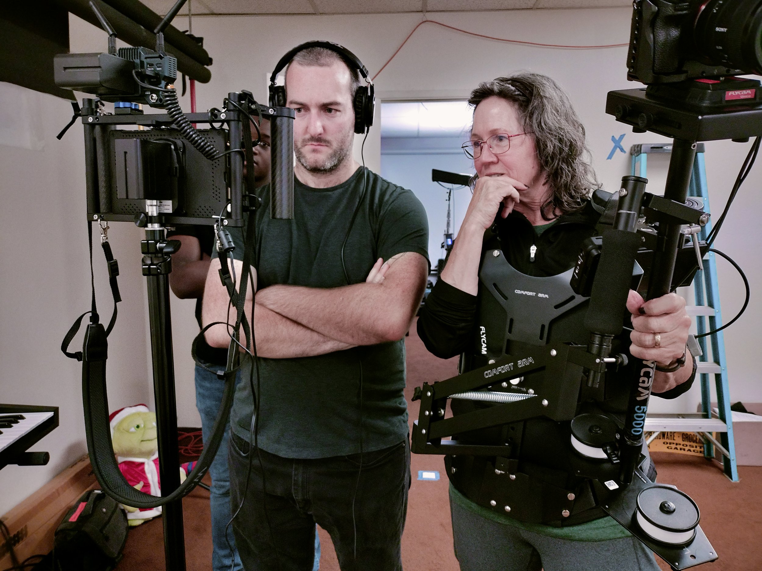 Four people in a room with camera equipment, two are in focus viewing a monitor, one is partially visible behind the monitors, and another person is in the background, all engaged in filming or monitoring a shoot.