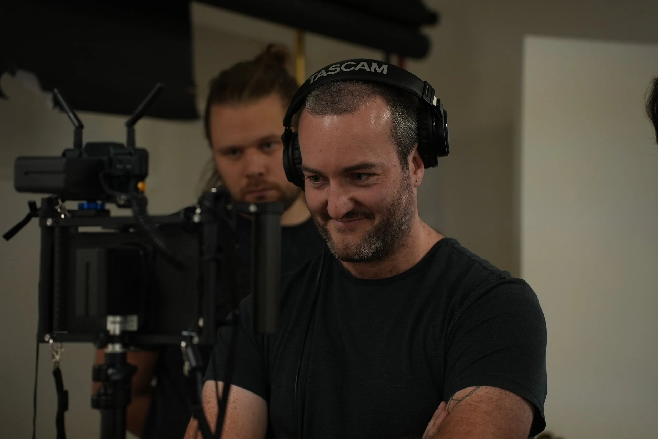 Two men working on a film set, one wearing headphones and operating a camera, the other observing in the background.