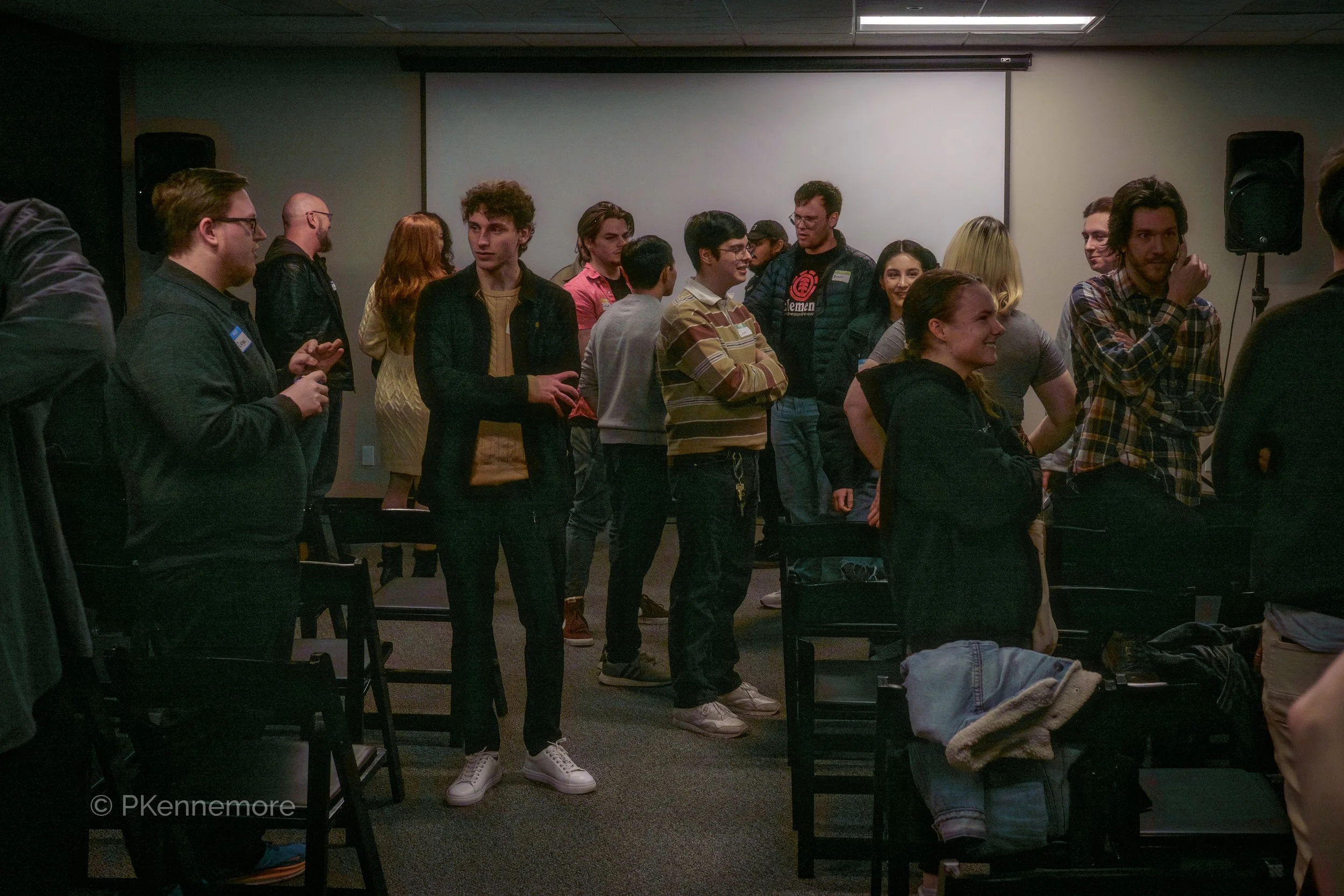 Group of people standing and talking in a dimly lit room with a projection screen on the wall.