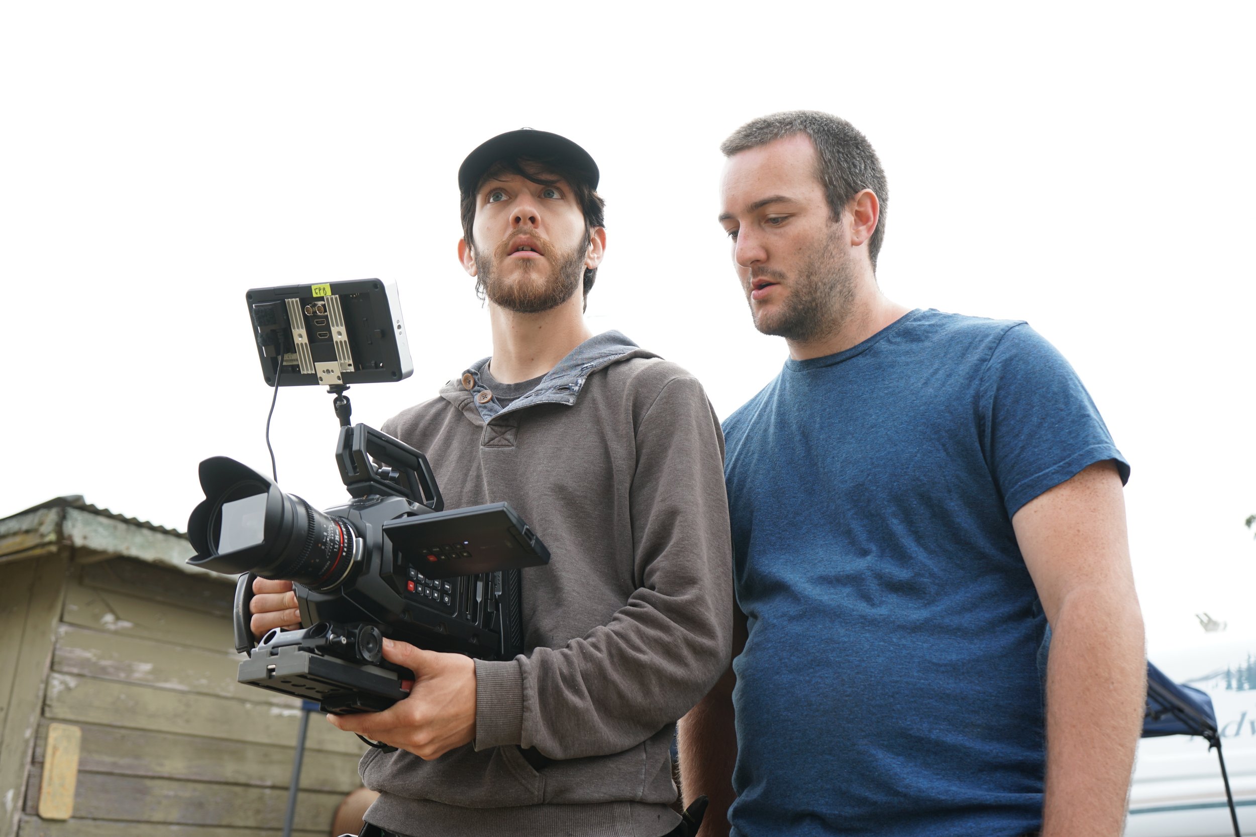 Two men operate a professional camera outdoors, with one man holding the camera and the other looking at it, against a cloudy sky and a wooden structure in the background.