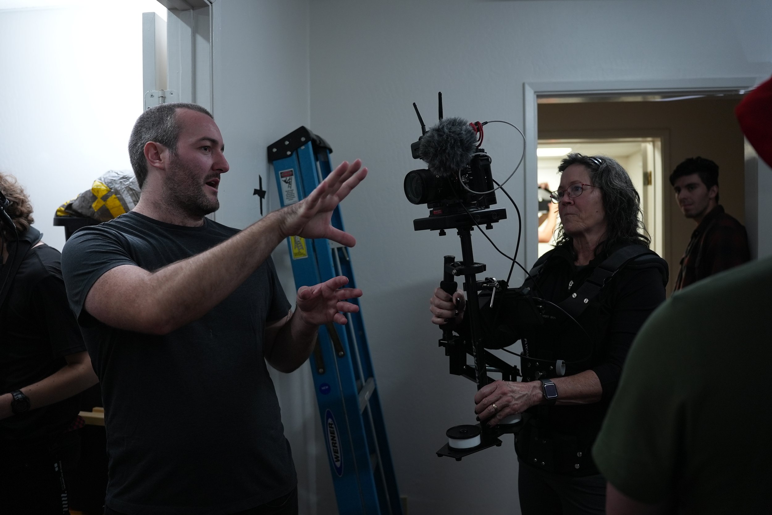 A man explaining something to a woman holding a professional camera rig during a film shoot, with a ladder and other crew members in the background.