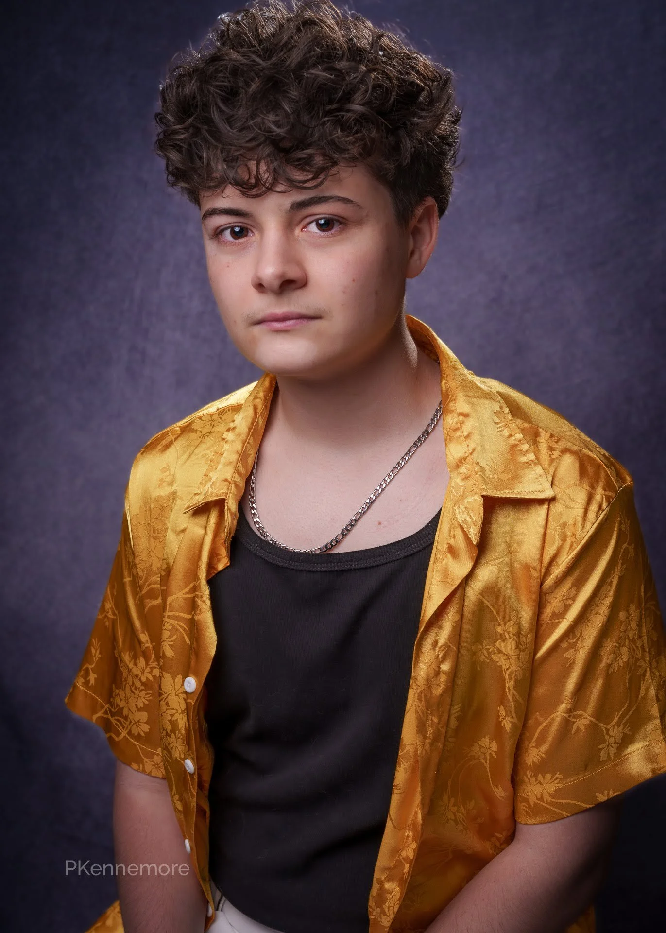 Portrait of a young man with curly brown hair, wearing a black shirt, a gold patterned short-sleeve button-up shirt, and layered silver chains, against a dark textured background.