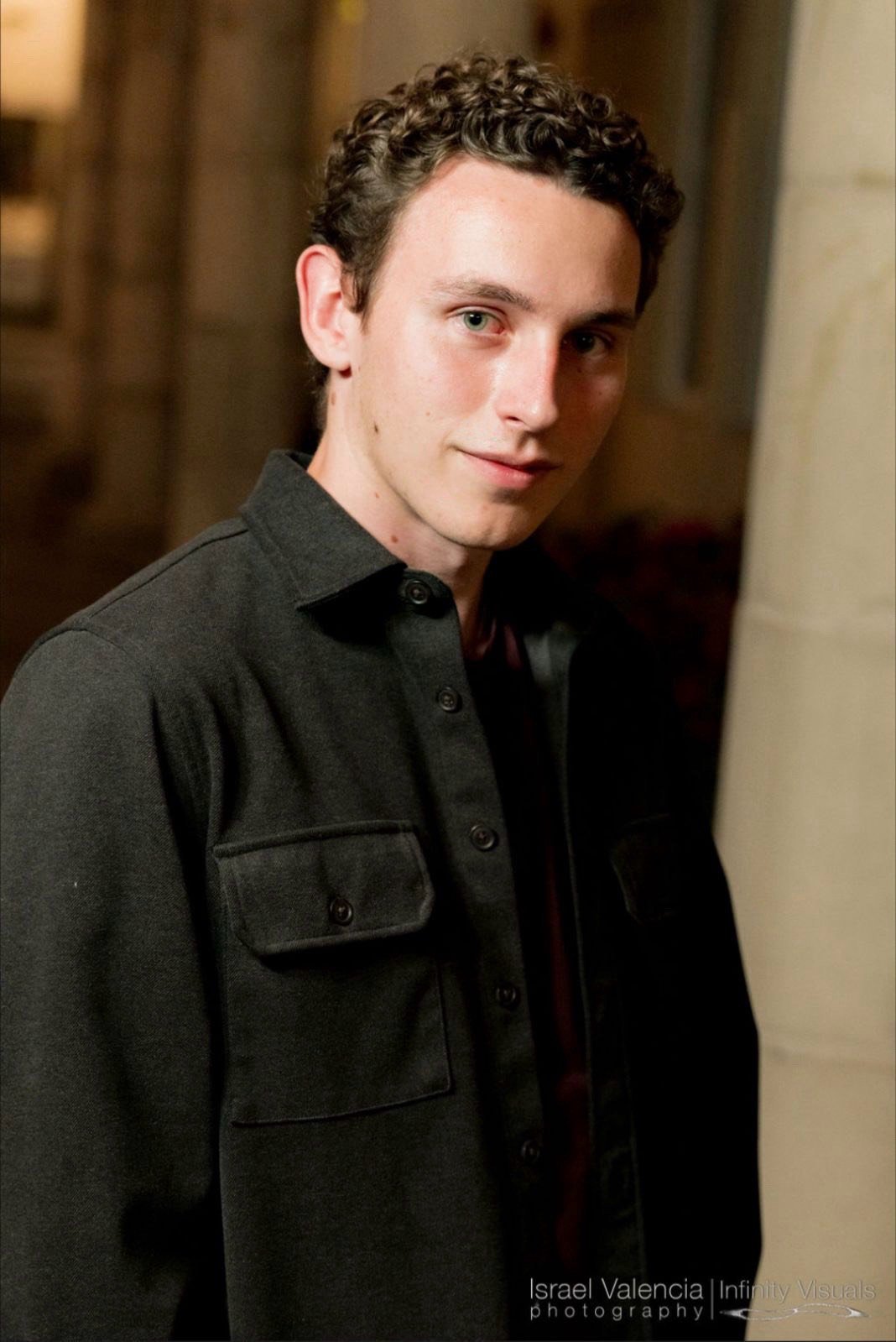 A young man with curly brown hair and blue eyes standing indoors near a stone wall, wearing a dark button-up shirt.