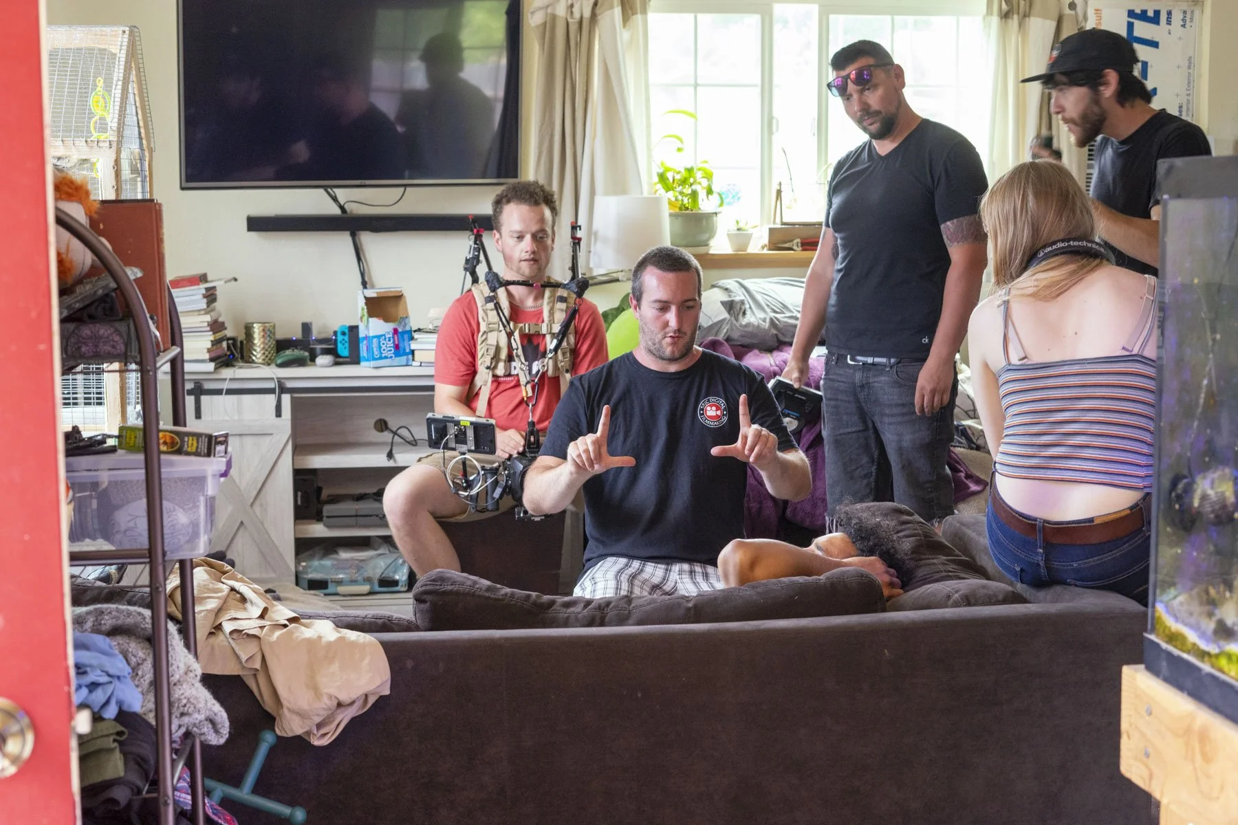 A film crew filming a scene in a cluttered living room with five people and various equipment, including a camera, lights, and wires, and a person lying on the couch.