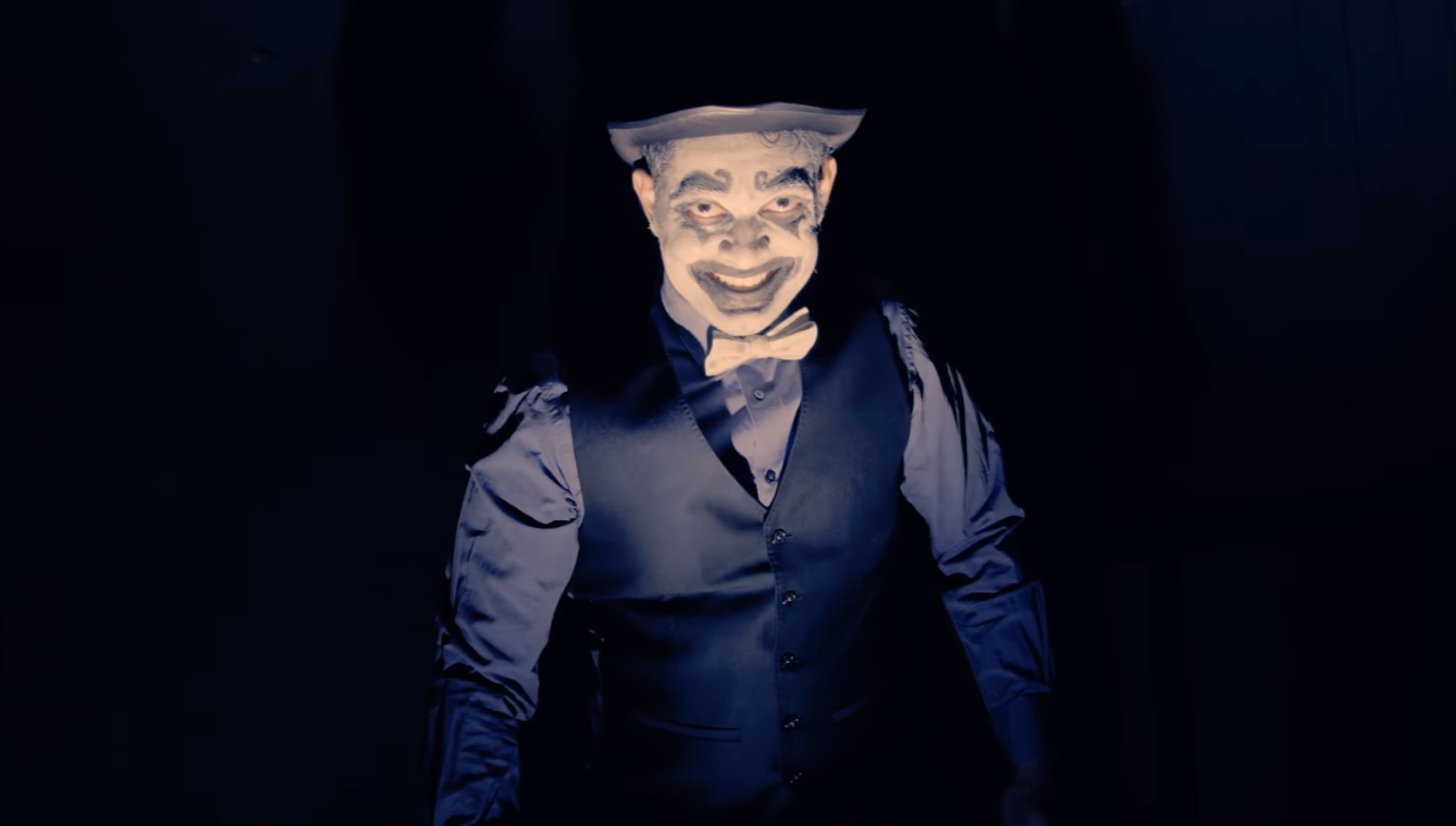Person dressed as a creepy clown with painted face, wearing a top hat, dark vest, and bow tie, standing in darkness.