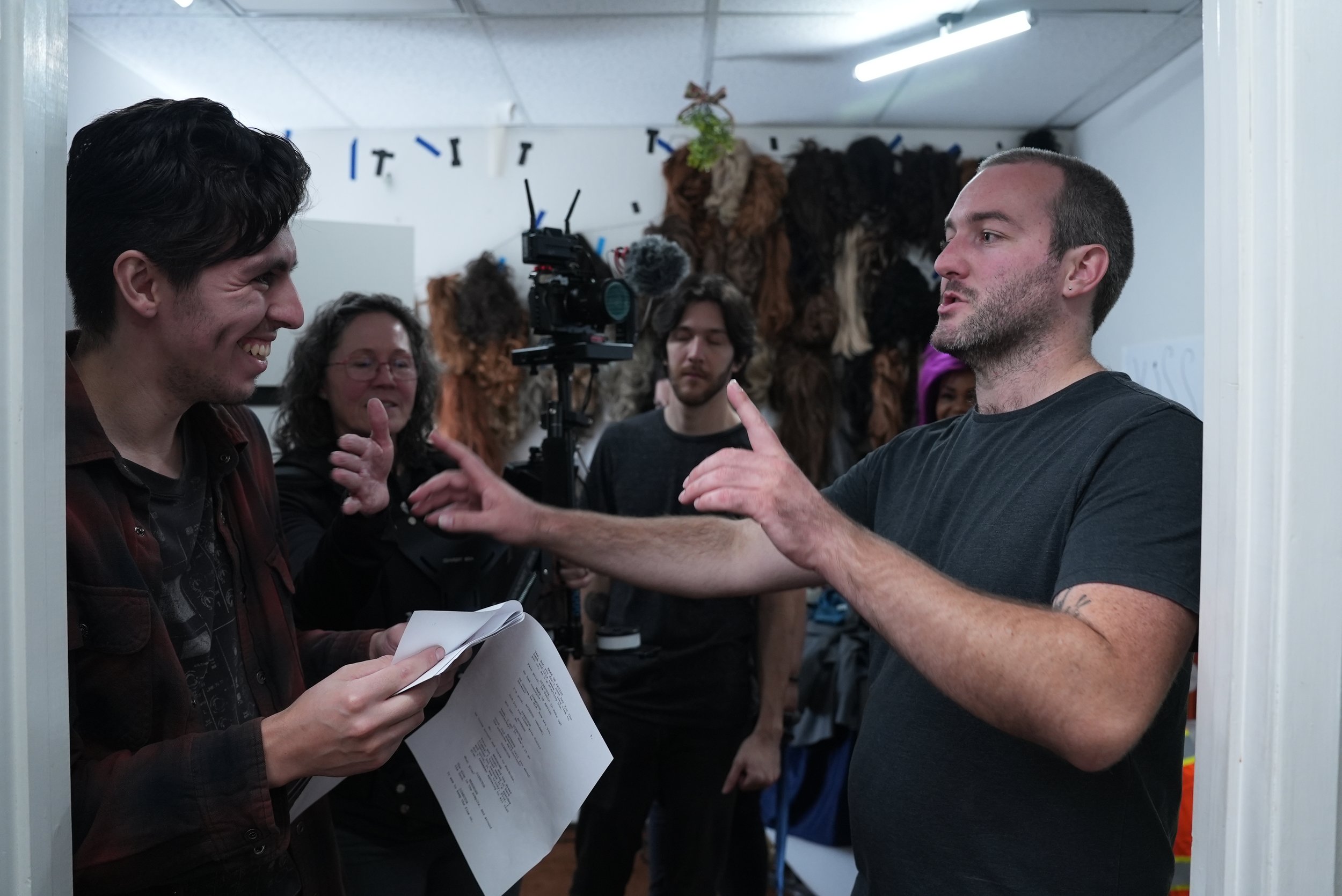 A group of people on a film or TV set, with one man in a black t-shirt talking to another man holding a script, while others watch, with costumes or props hanging in the background.