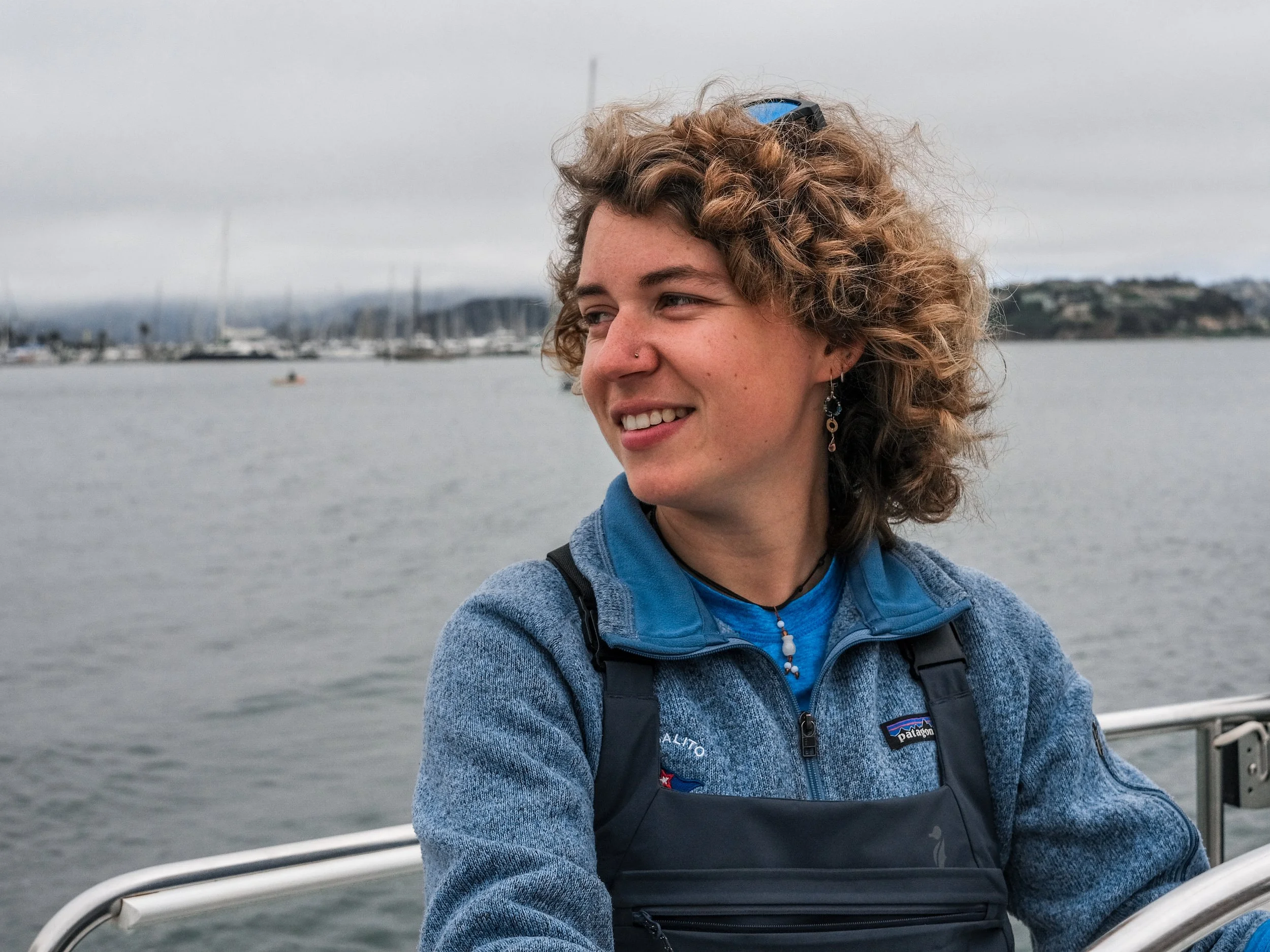A woman with curly hair smiling on a boat with a marina and sailboats in the background under cloudy skies.