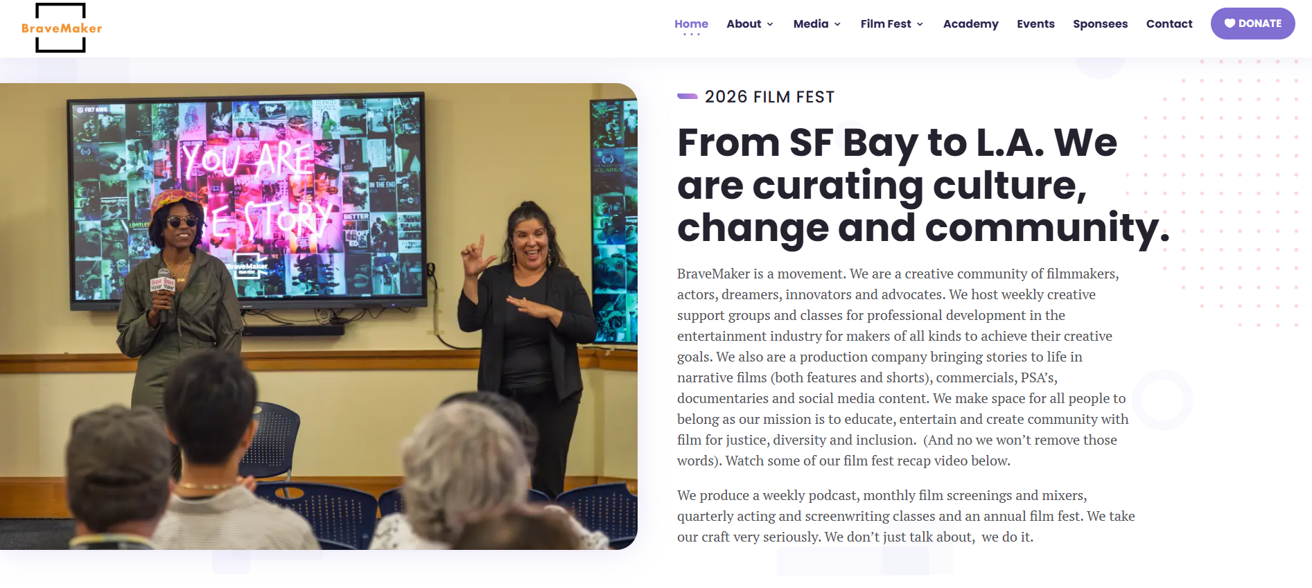 Two women speaking at a community event, one with sunglasses and a hat holding a microphone, the other with long curly hair gesturing as she speaks, in front of a large screen displaying colorful images and a neon sign that reads 'YOU ARE THE STORY'. Audience members are seated facing the speakers.