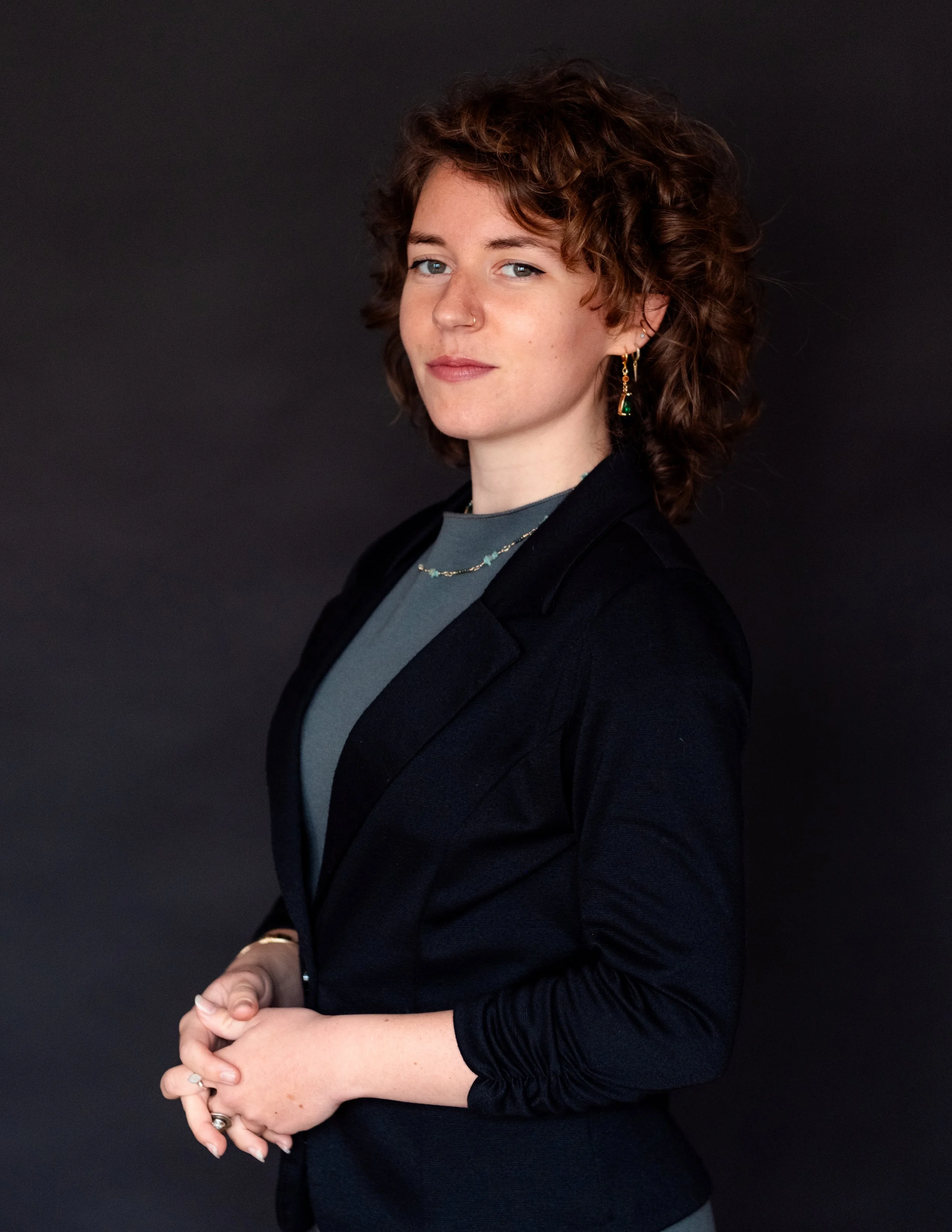 A woman with curly brown hair wearing a black blazer, gray shirt, earrings, and jewelry posed against a dark background.