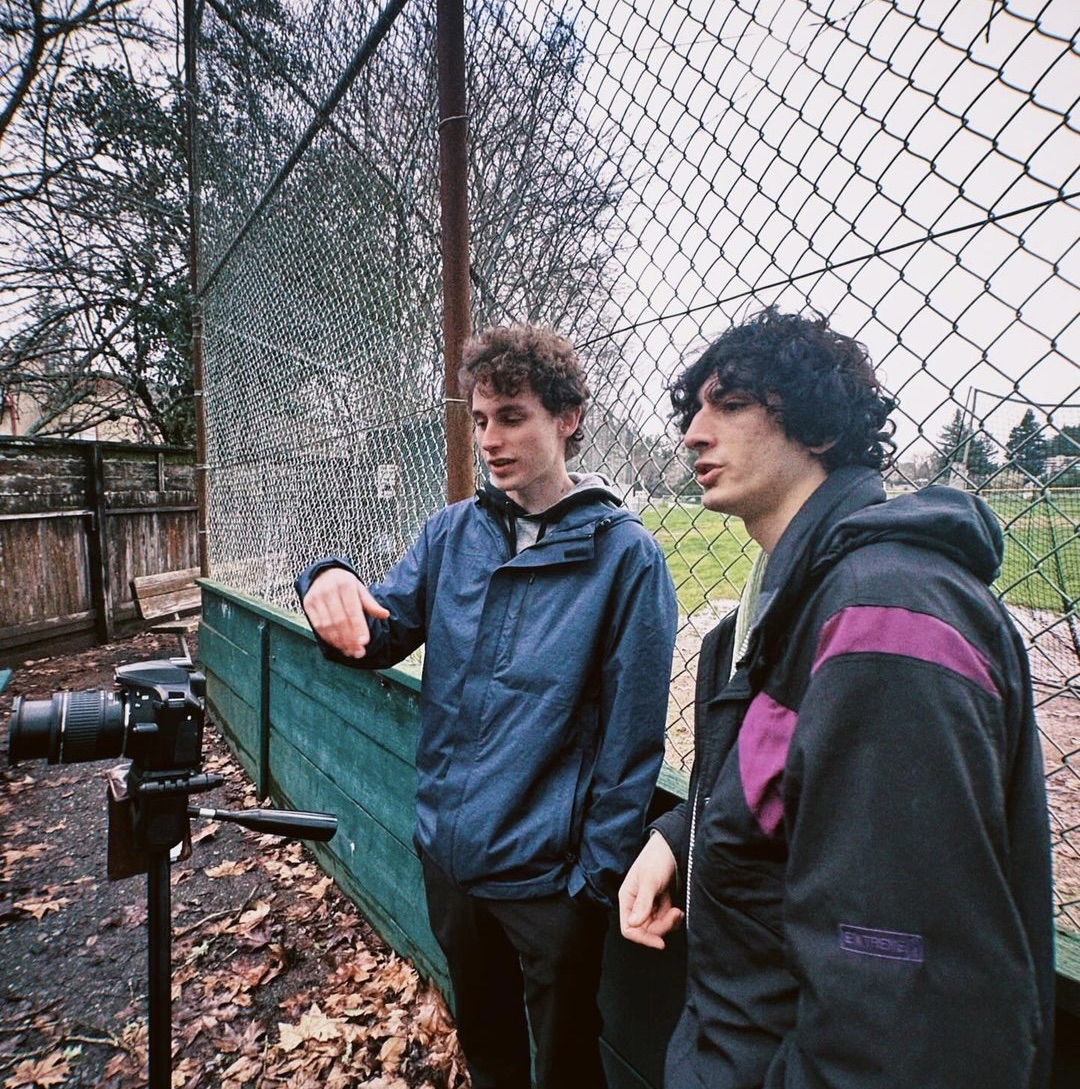 Two young men talking by a camera on a tripod next to a chain-link fence on a cloudy day, with fallen leaves on the ground.