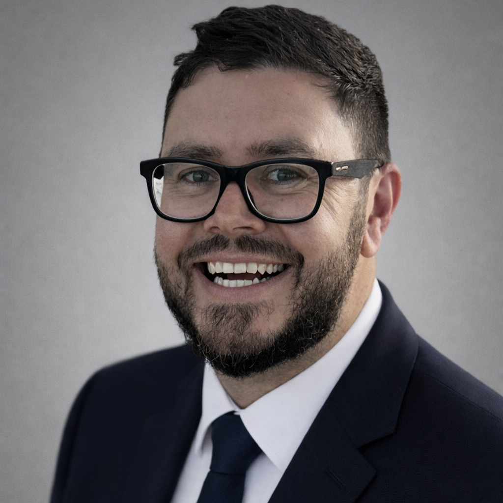 A man with dark hair, glasses, and a beard, smiling and dressed in a suit against a plain gray background.
