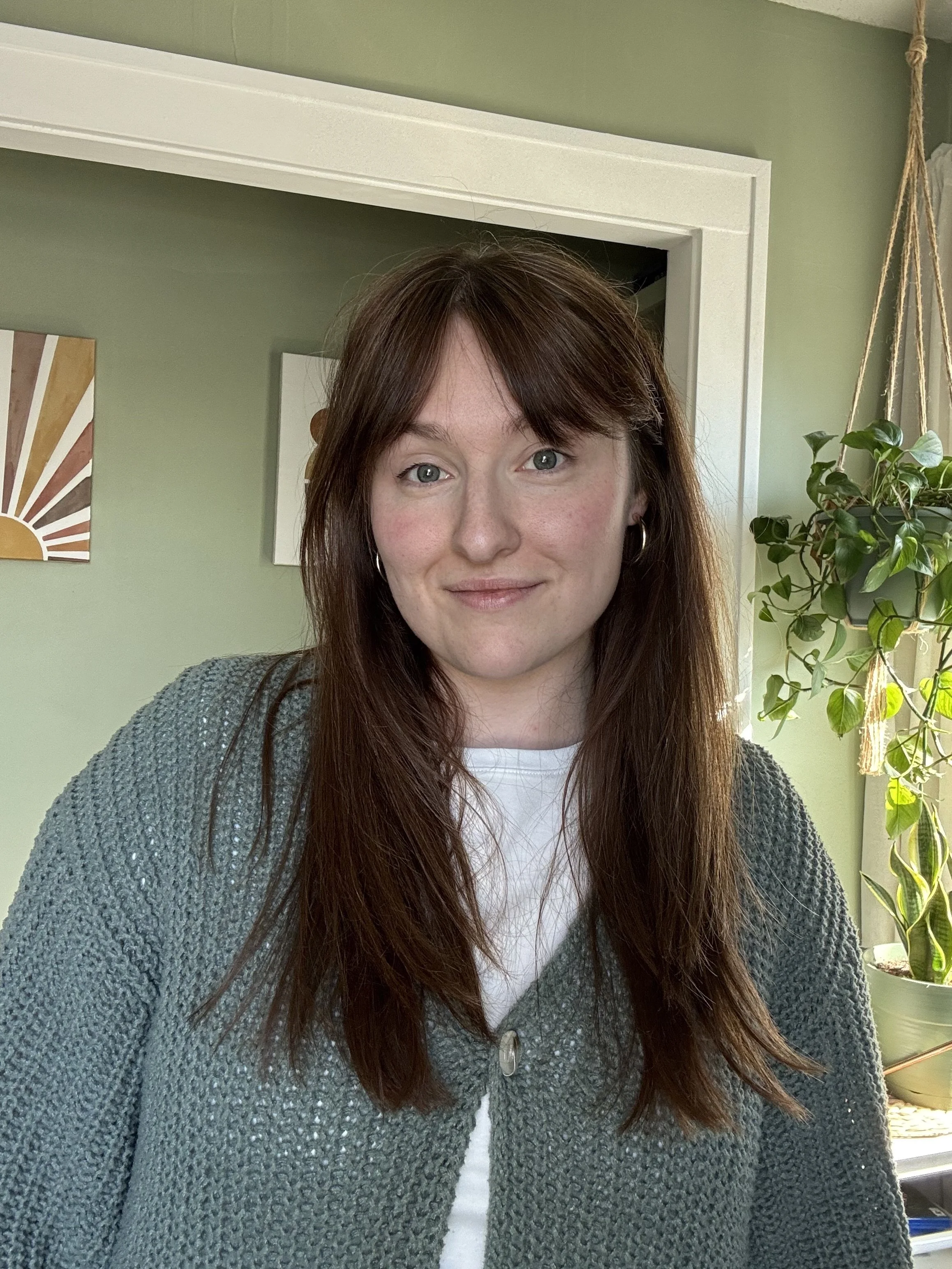 A woman with long brown hair and blue eyes smiling in a room with green walls, a window with sunlight, hanging plants, and framed artwork.