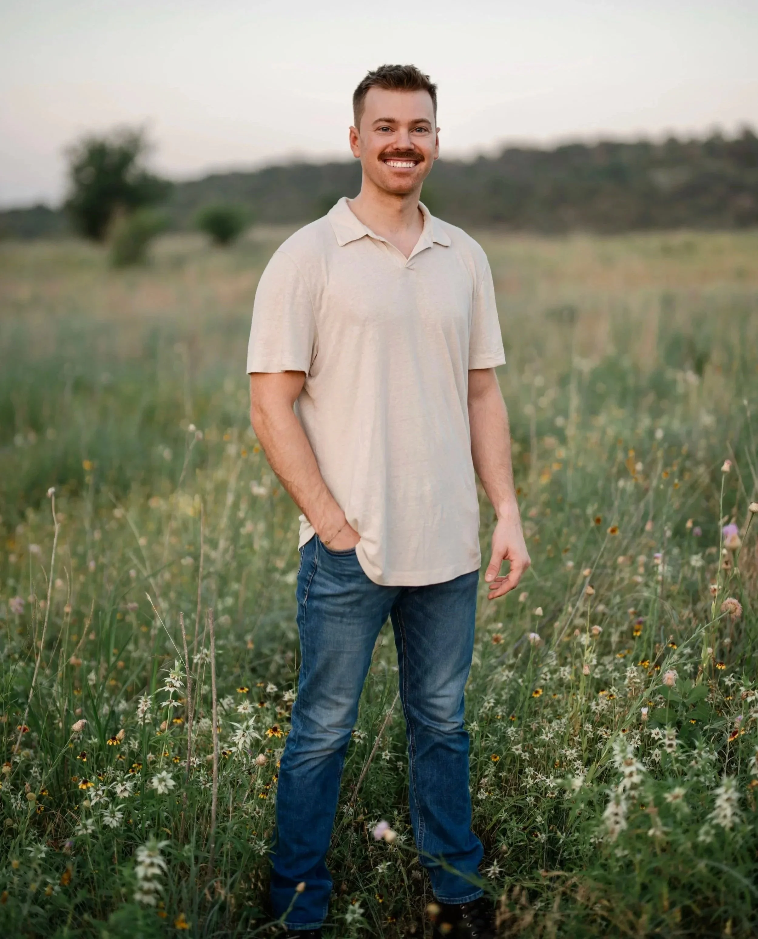 A young man with short brown hair, a beard, and wearing a light beige polo shirt and blue jeans, standing in a grassy field with wildflowers, smiling at the camera during the daytime.