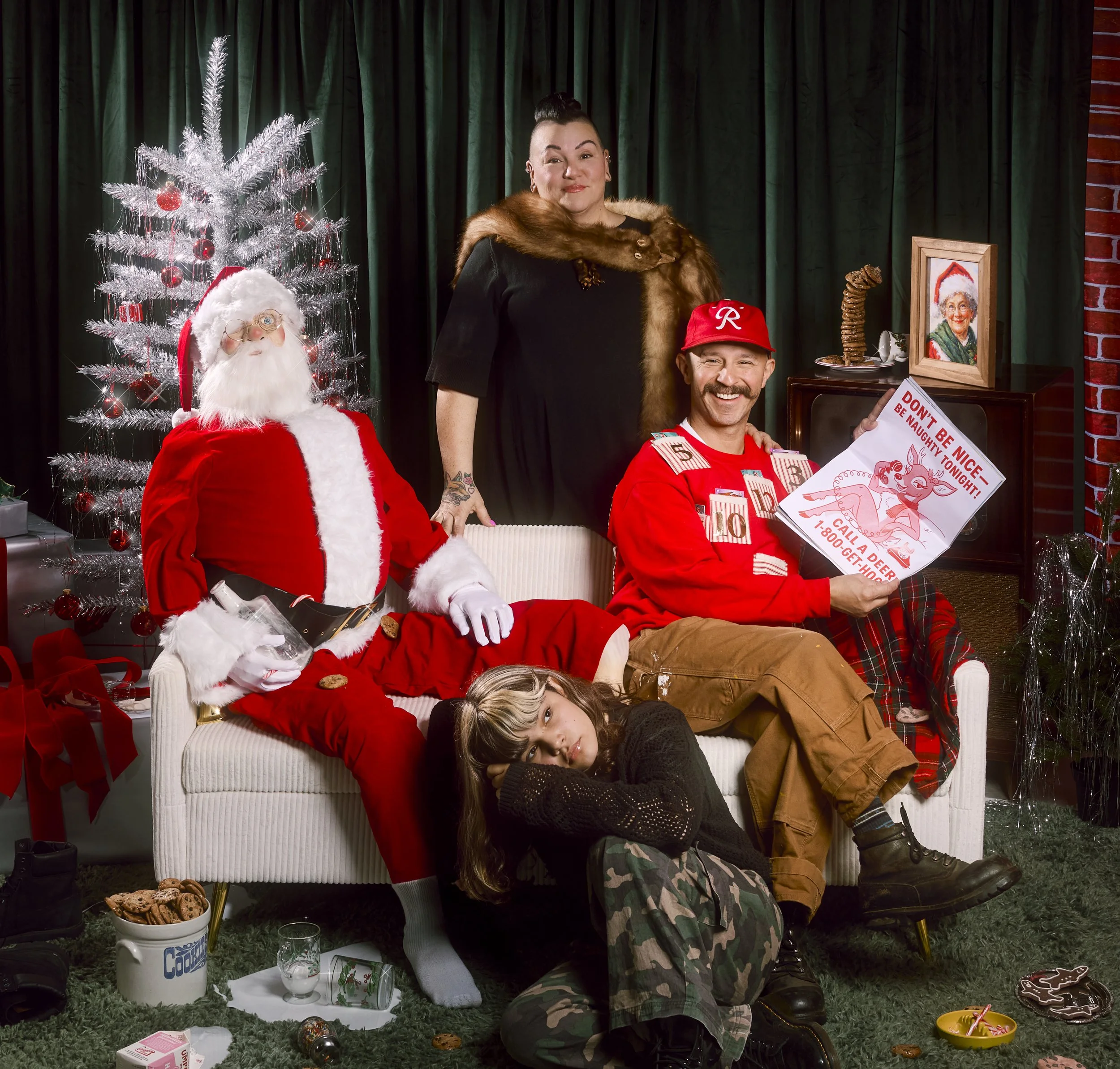 A woman and two children with Santa Claus and a man dressed in Christmas-themed clothing in a festive room with a white Christmas tree, decorations, and Christmas snacks.