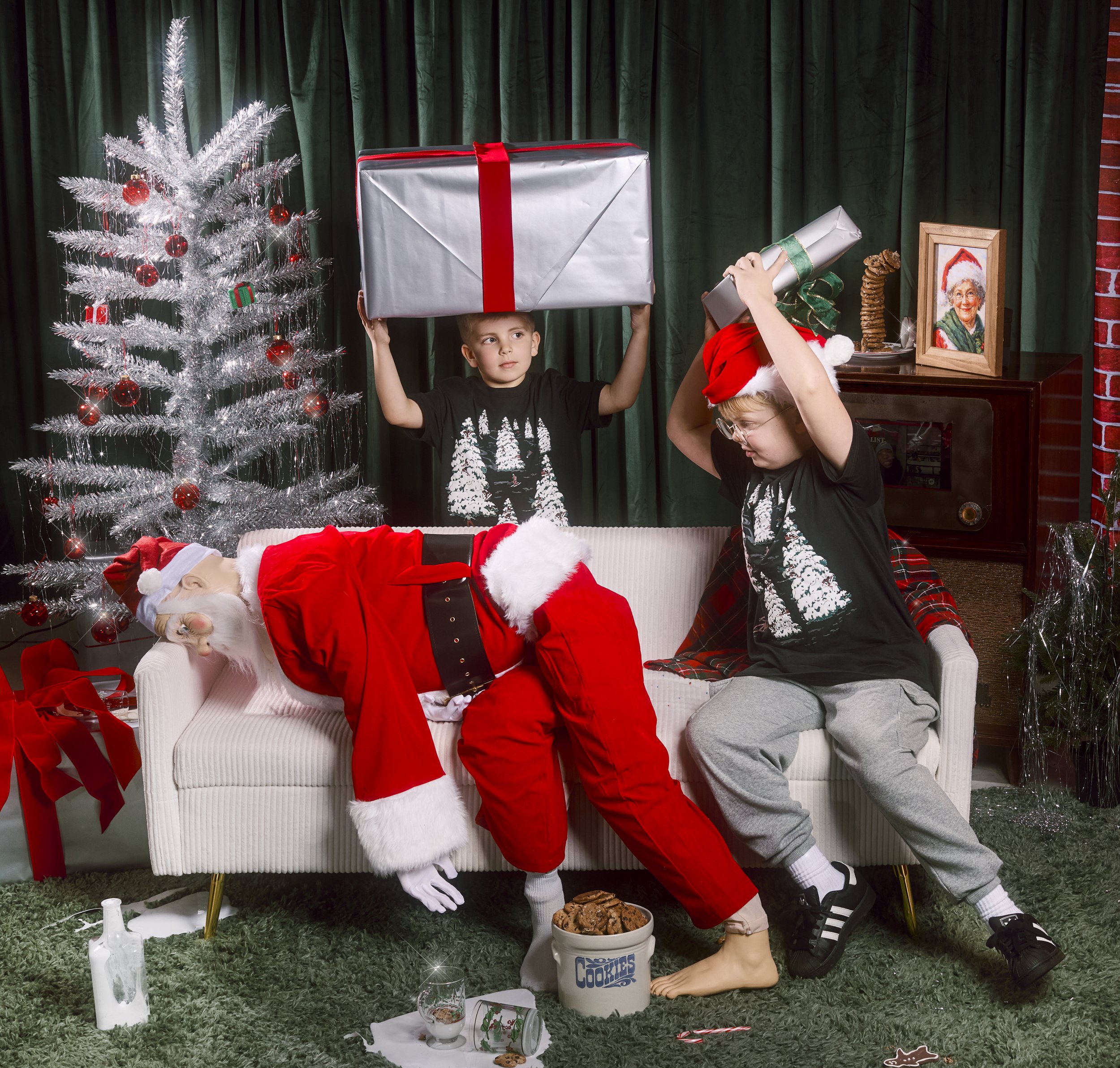 Children in Christmas pajamas and Santa costume on a sofa with Christmas decorations, one child with a Santa hat holding a large gift above his head, another with a Santa hat placing a sprig on Santa's head, Christmas tree with red ornaments, present