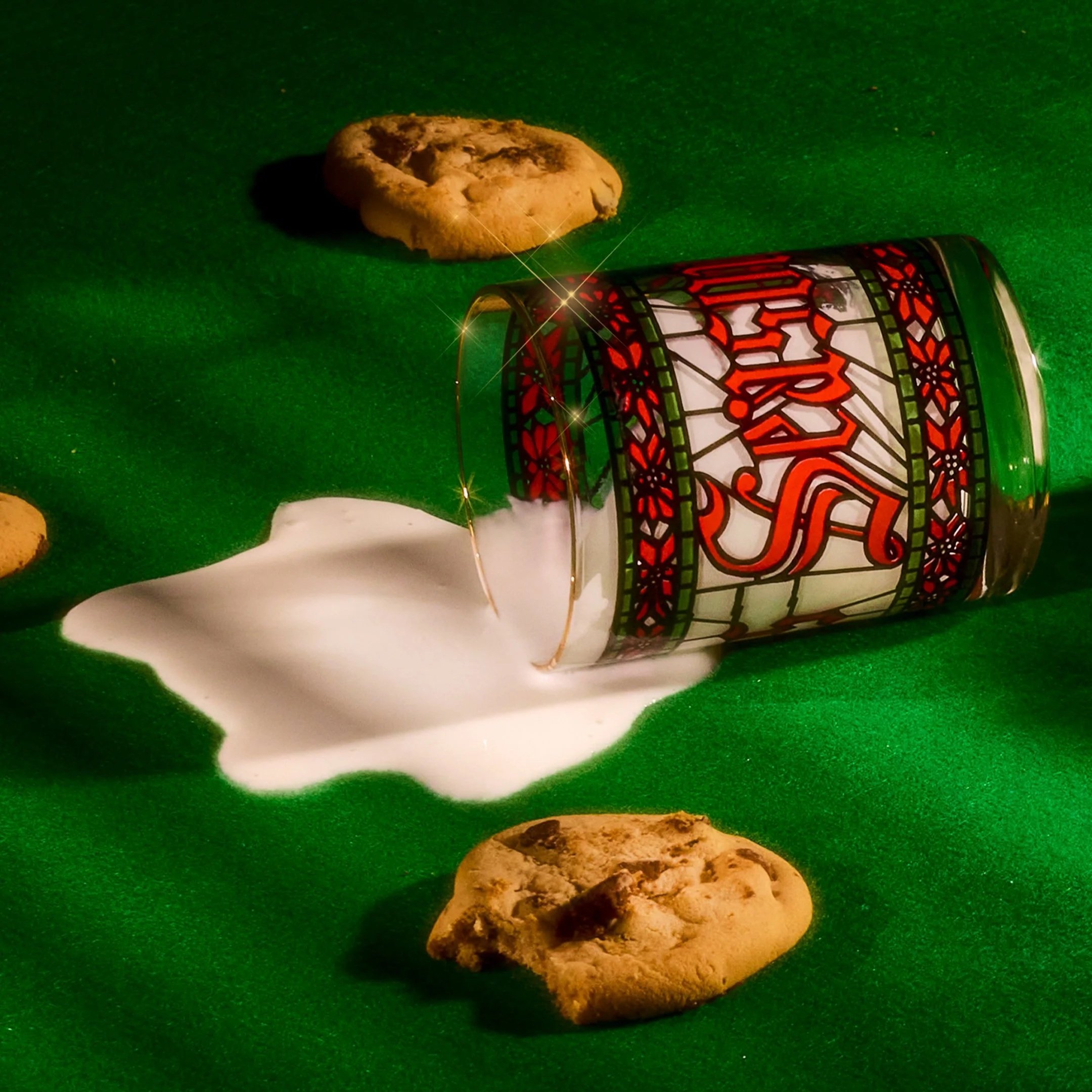 An overturned glass decorated with red and green stained glass stained glass pattern lies on green fabric, spilling milk or cream, with chocolate chip cookies nearby.