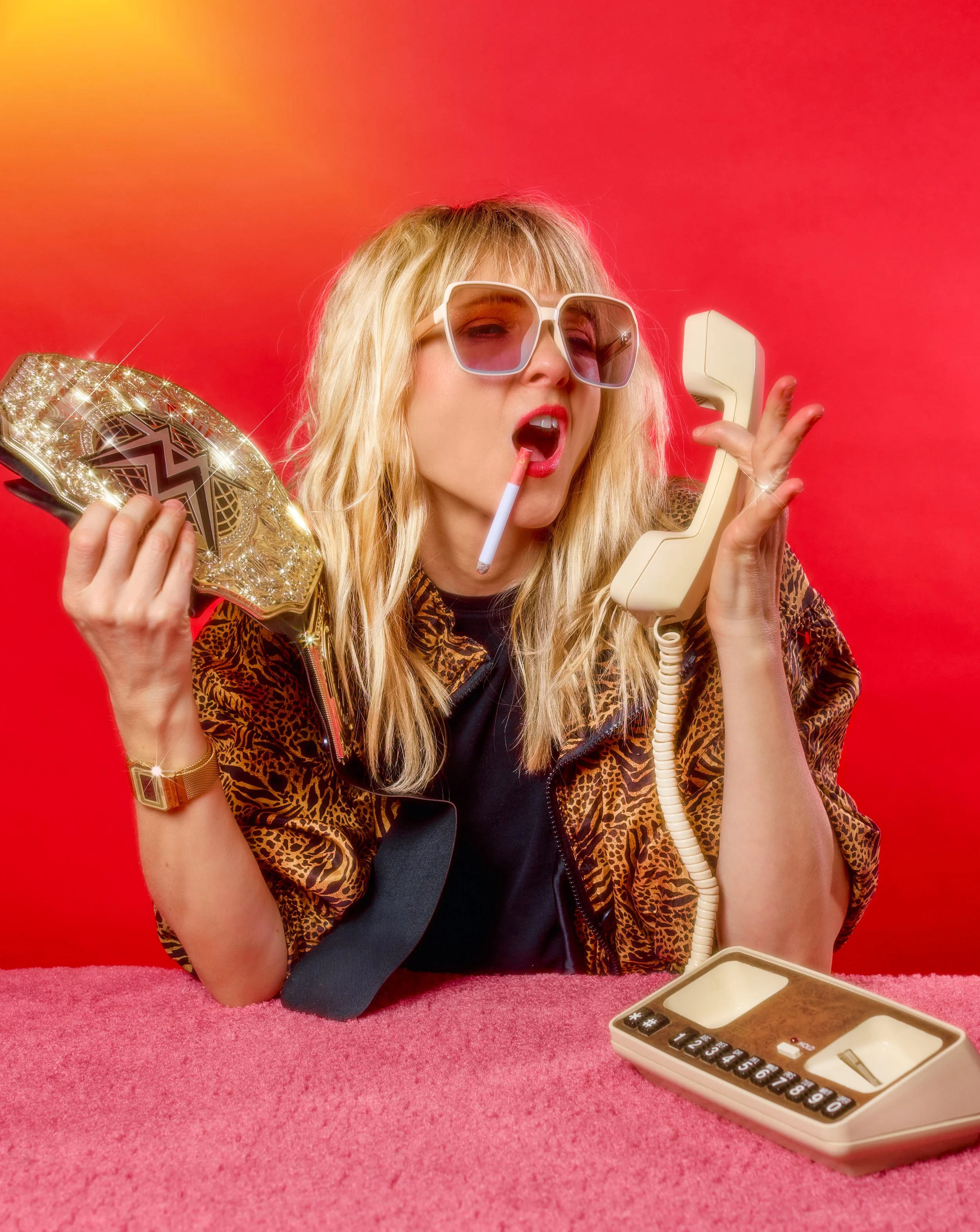 A woman with blonde hair, oversized sunglasses, and a cigarette in her mouth, wearing a leopard print jacket, holds a wrestling championship belt in one hand and an old-fashioned rotary phone in the other, sitting at a pink surface with a red background.
