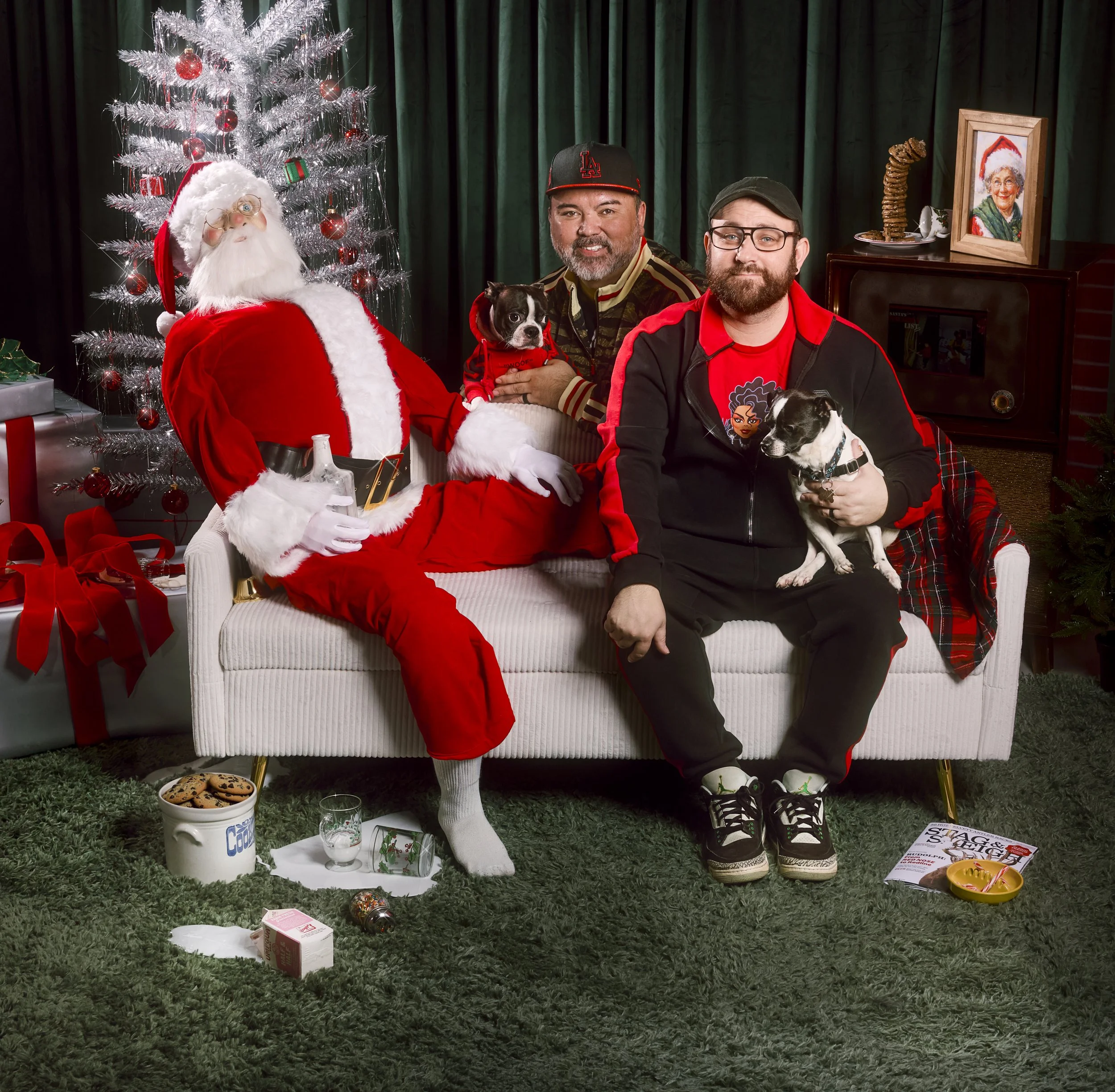 Two men sitting on a white couch with two dogs, posing with a Santa Claus figure during Christmas. The setting includes a Christmas tree with red ornaments, gifts, and festive decorations.
