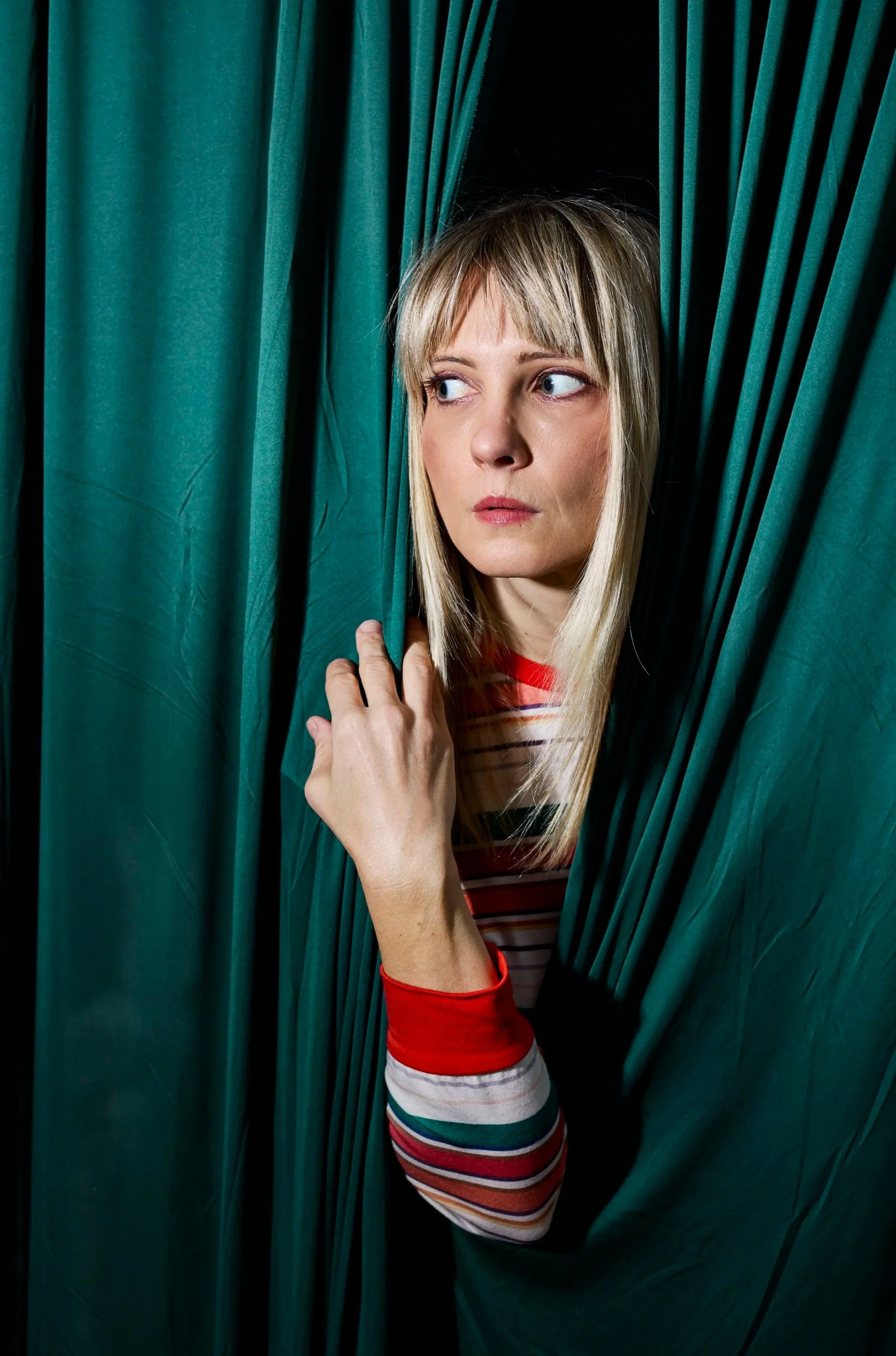 A woman with blonde hair and light skin peeks out from behind green theater curtains, wearing a striped shirt with red, white, and black stripes.