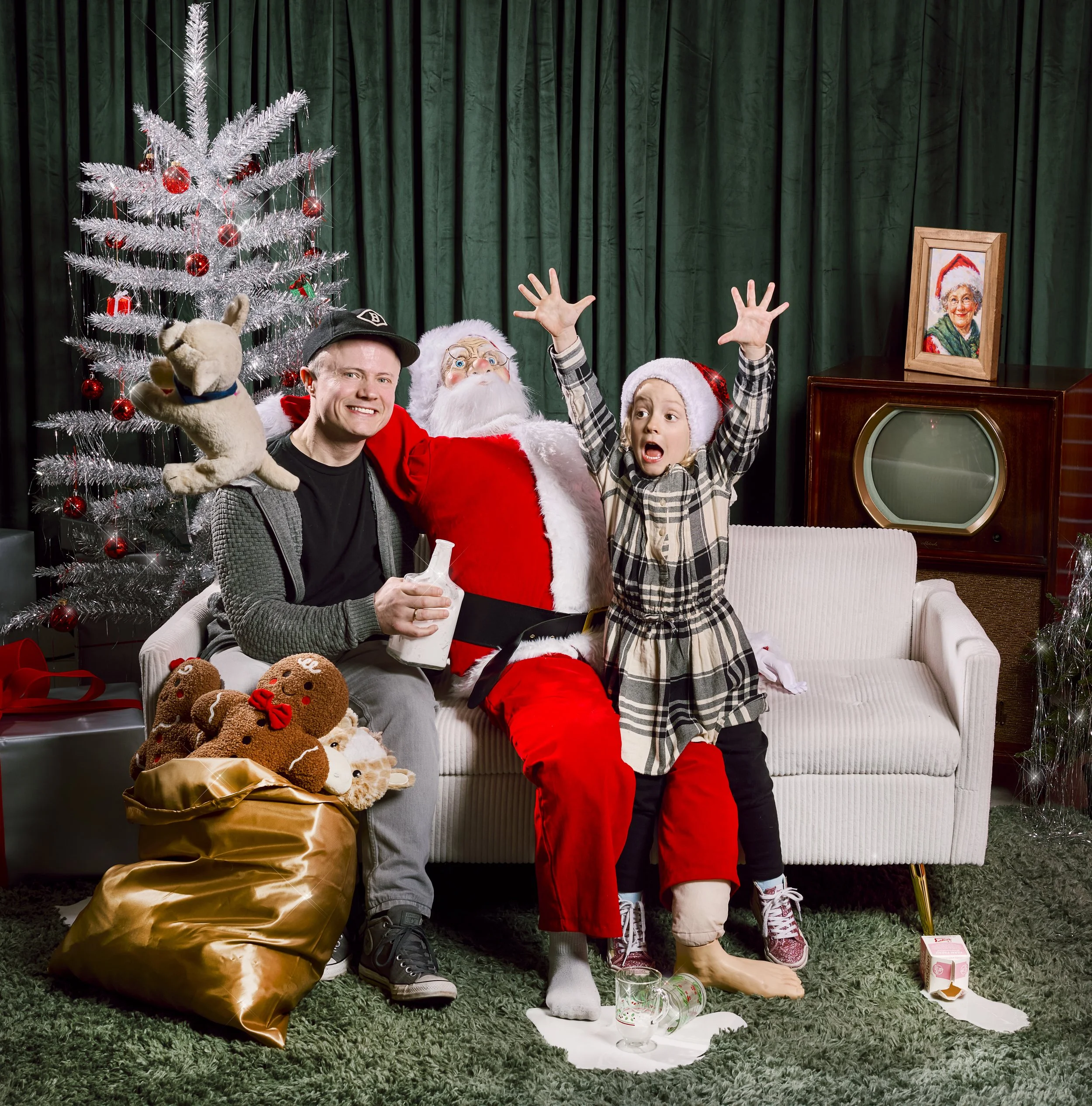 A man, Santa Claus, and a young girl sitting on a white sofa in a festive holiday scene. The man is holding a plush reindeer toy, Santa has a glass of milk, and the girl is raising her hands excitedly. A decorated Christmas tree with red ornaments is