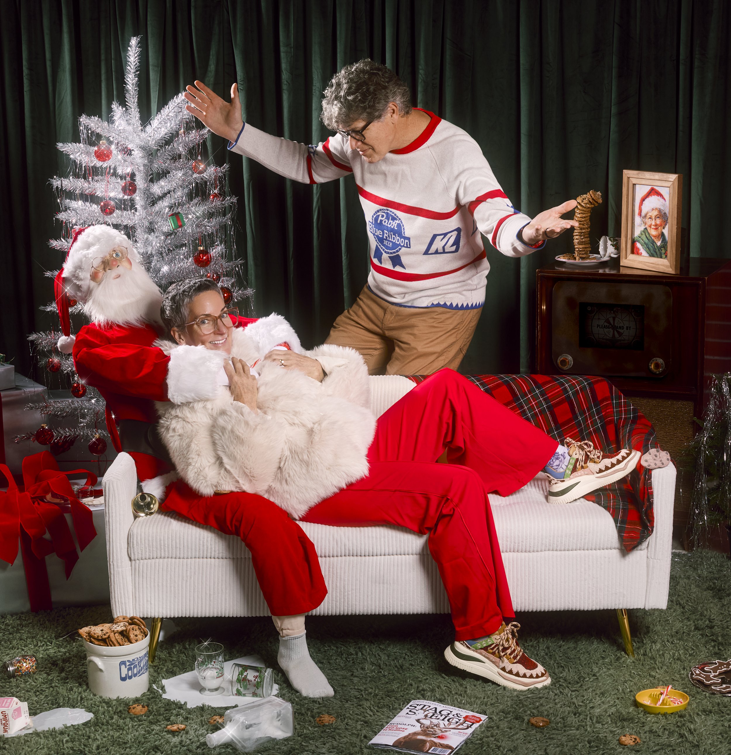 A woman dressed as Santa Claus lying on a white couch with a woman wearing glasses smiling, a woman dressed as Santa Claus, and a man standing on the couch with arms outstretched in a festive living room with a Christmas tree, presents, and holiday d