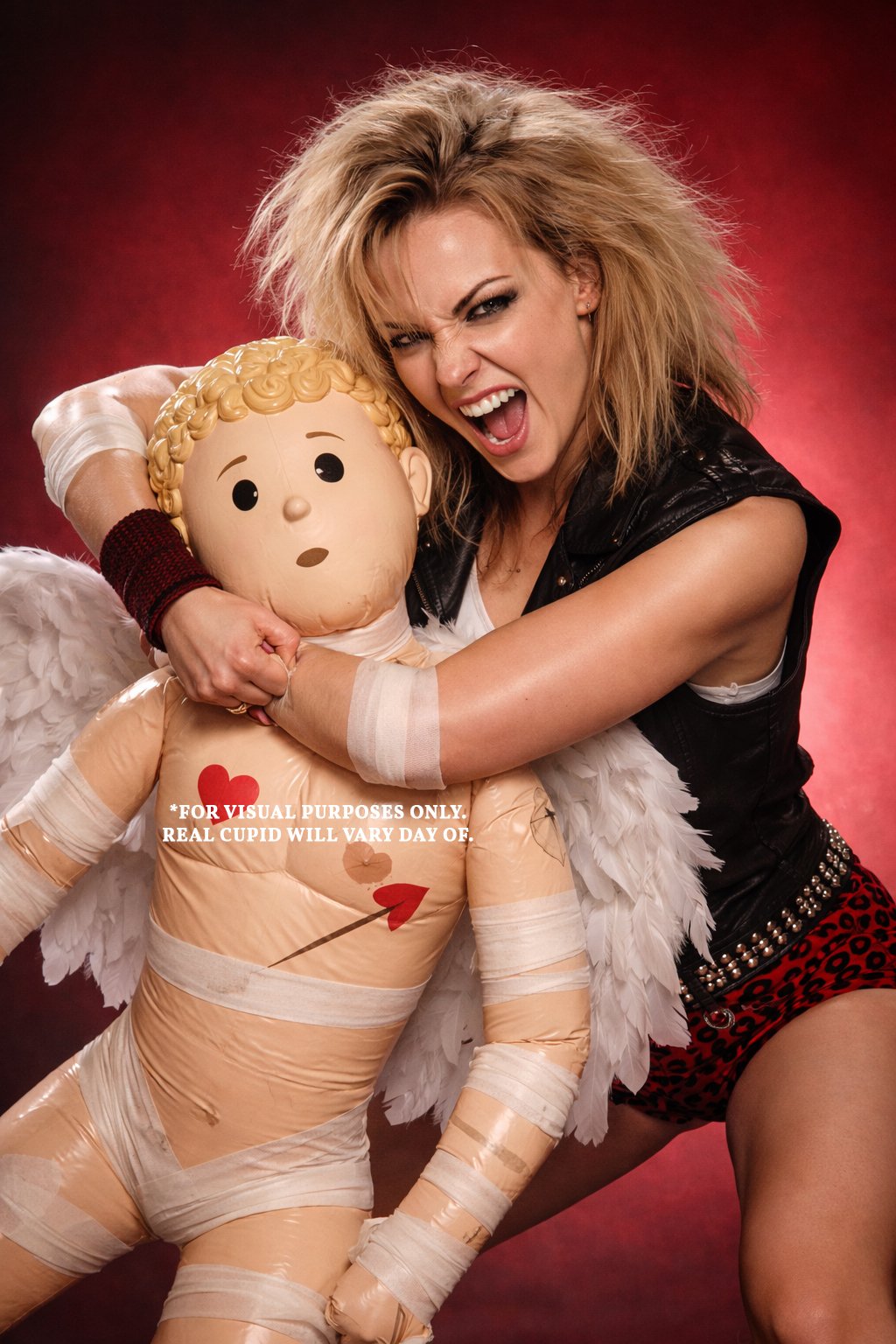 A woman with wild blonde hair, wearing a black leather vest and red leopard print shorts, hugs a large inflatable Cupid doll with wings, red heart markings, and playful facial expression against a red background.