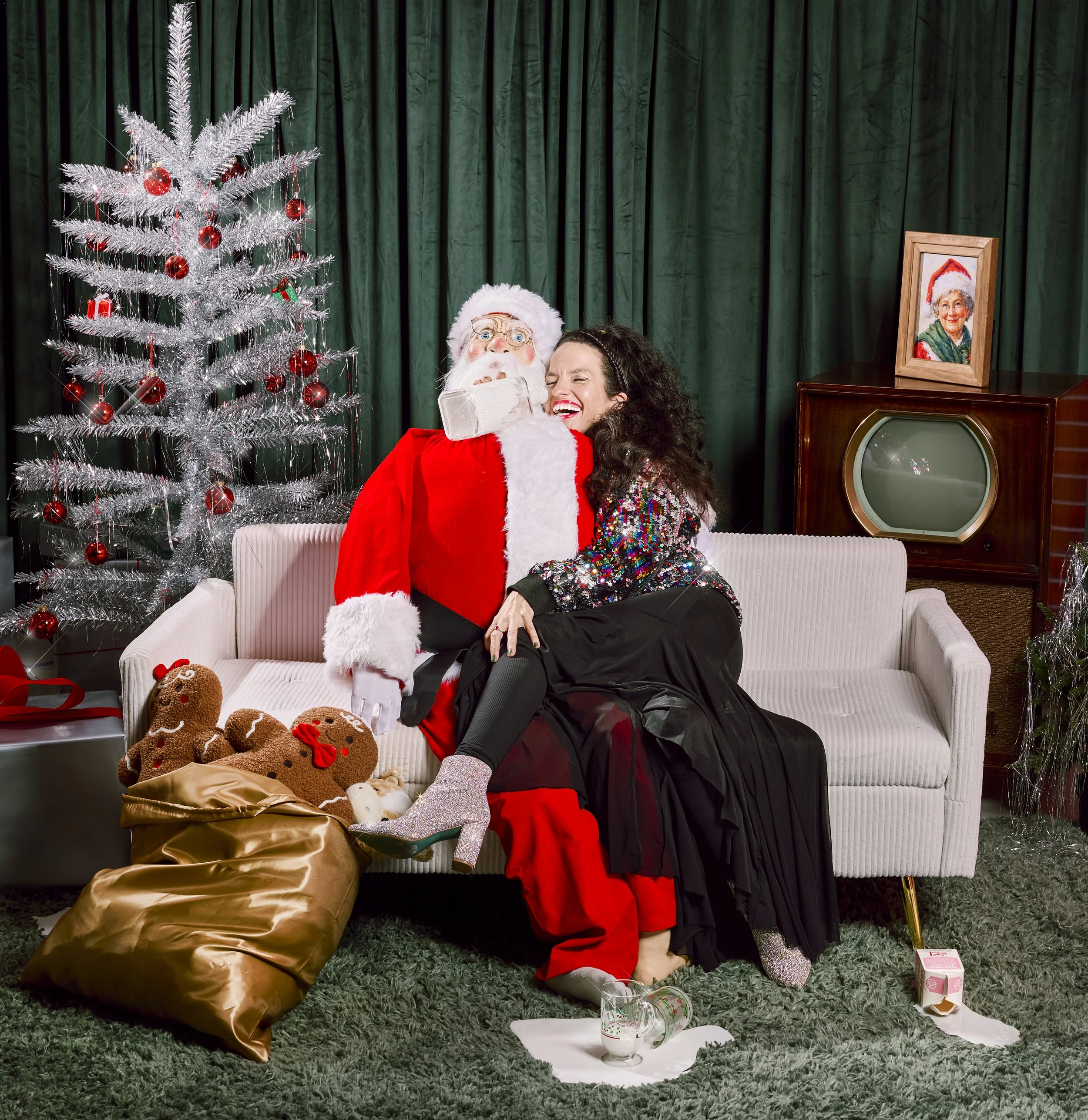 A woman in a sparkly top and black skirt sitting on Santa Claus's lap, laughing joyfully. Santa is dressed in a traditional red suit with white fur trim, in front of a decorated Christmas tree with red ornaments and tinsel. There are gingerbread cookies on a gold bag on the couch. The scene is festive with a vintage TV and a framed Christmas portrait in the background.