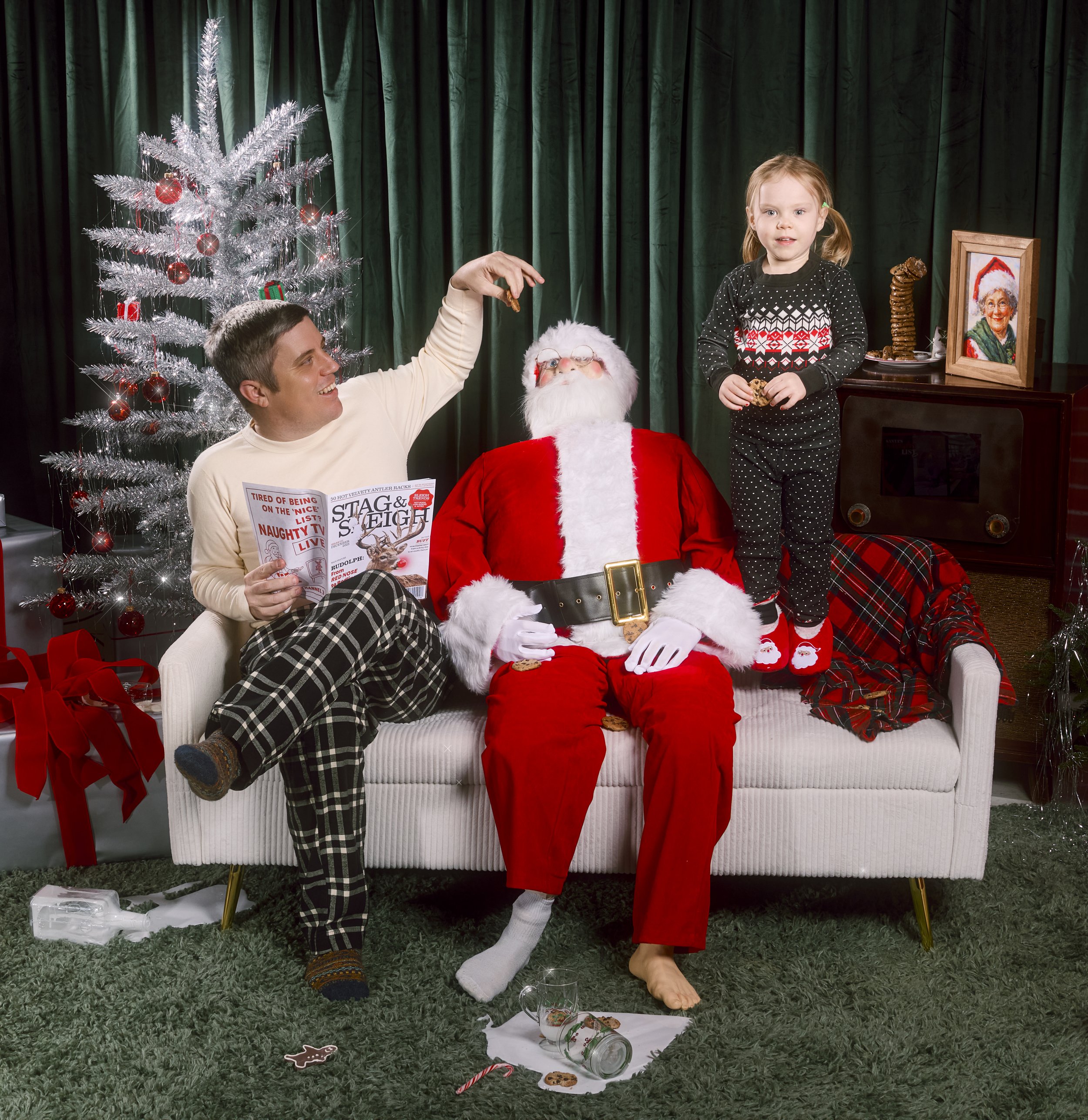 A family with a man, a young girl, and Santa Claus sitting on a sofa in a Christmas scene. The man is handing a cookie to Santa, who is seated in the middle. A Christmas tree decorated with red ornaments is in the background. The young girl is standi