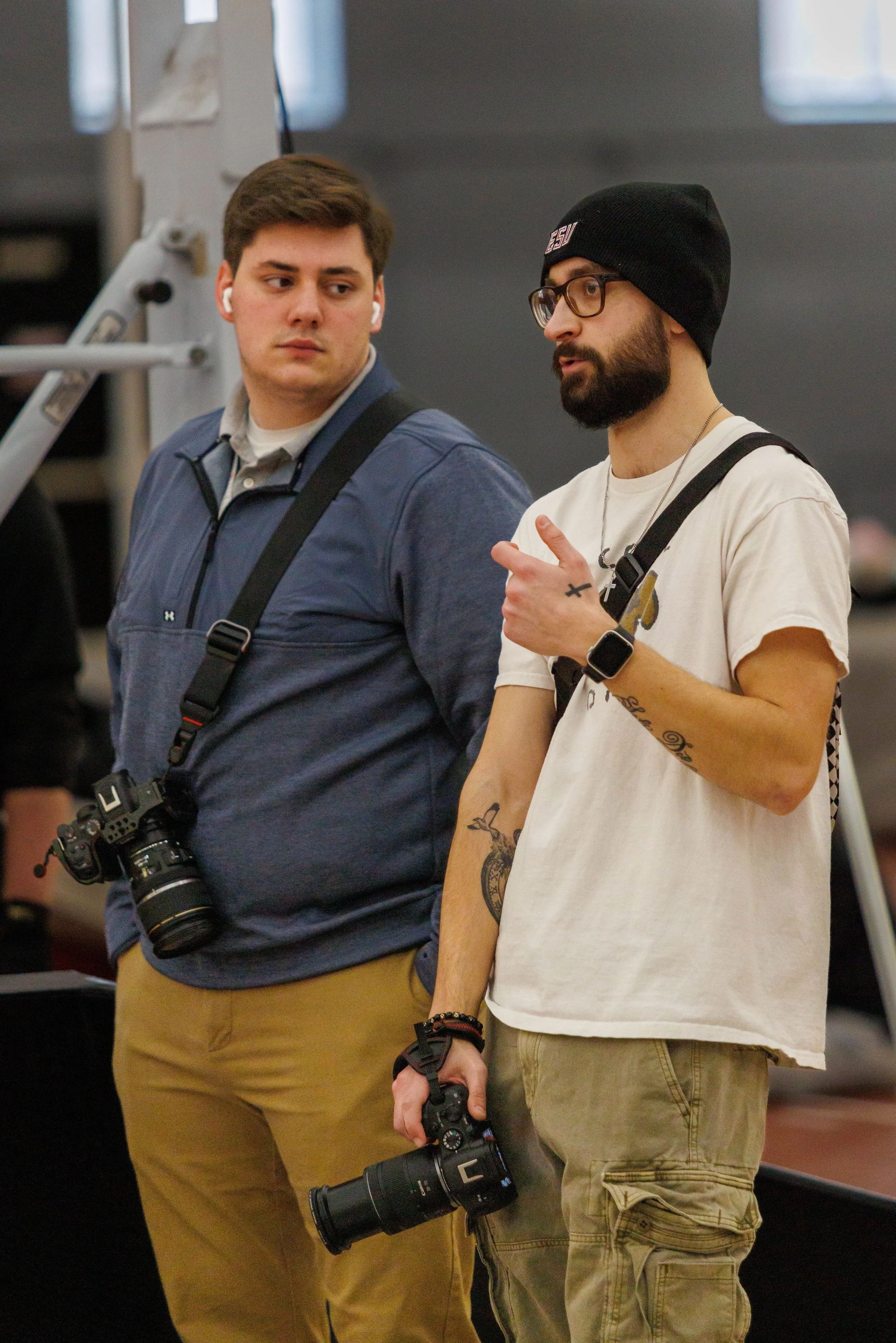 Two young men standing and talking, both holding cameras, one wearing glasses and a black beanie, the other with earbuds, in an indoor setting.