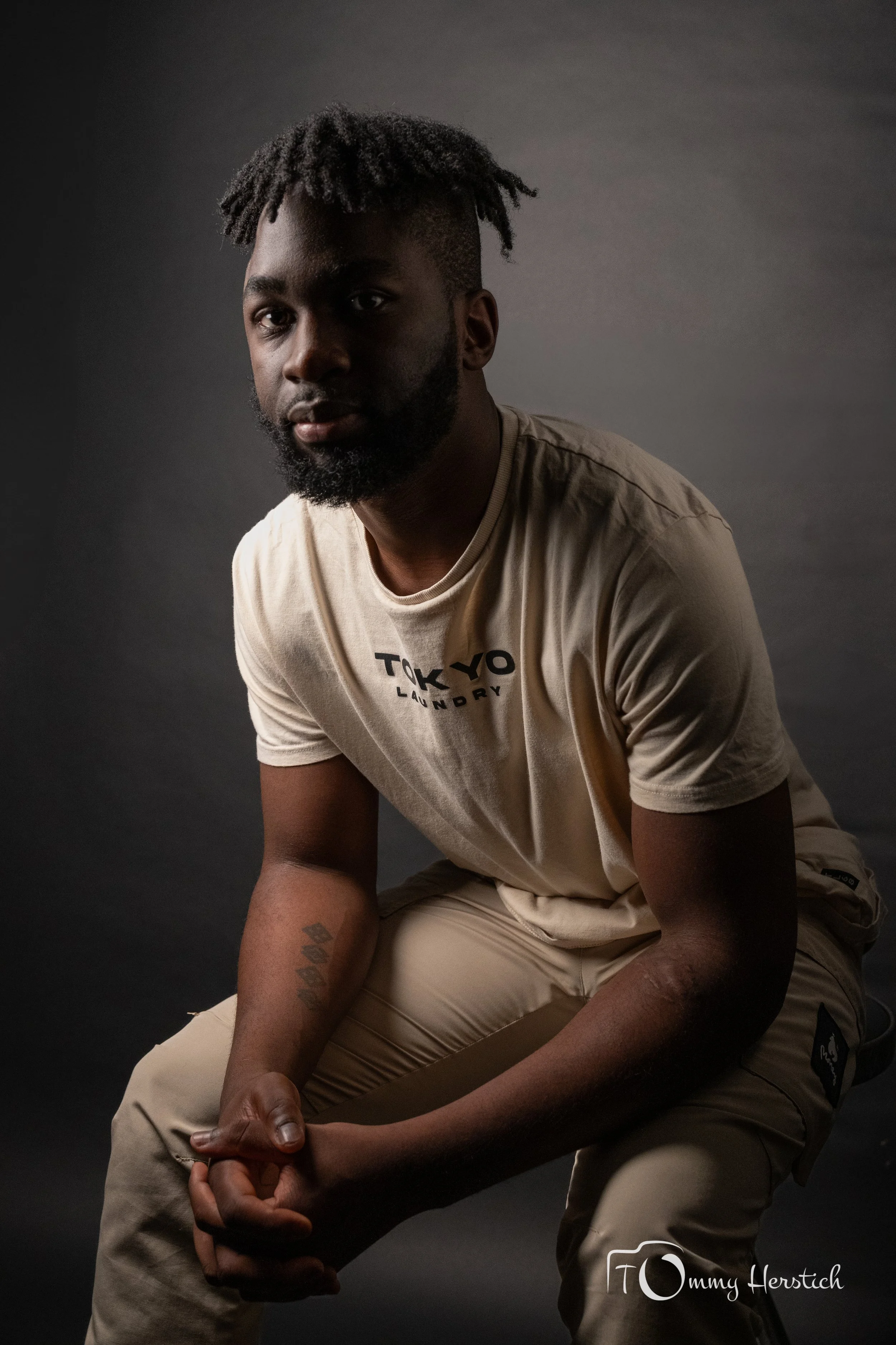 A young man with dreadlocks and a beard, wearing a beige T-shirt and beige pants, sitting against a dark background, looking directly at the camera.
