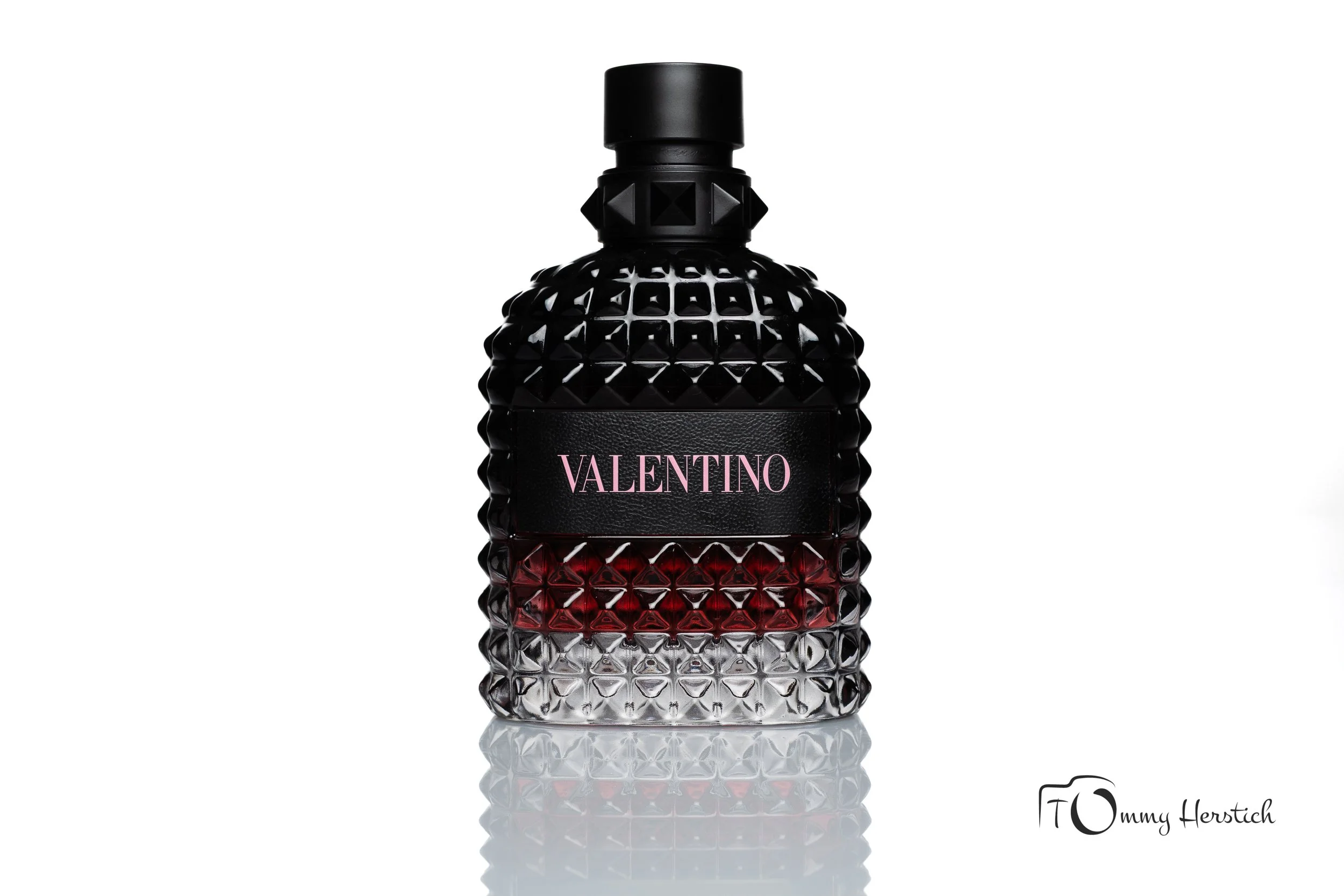 A black, textured perfume bottle with a diamond pattern, featuring a black cap and a prominent pink Valentino label, set against a white background with a slight reflection visible at the base.
