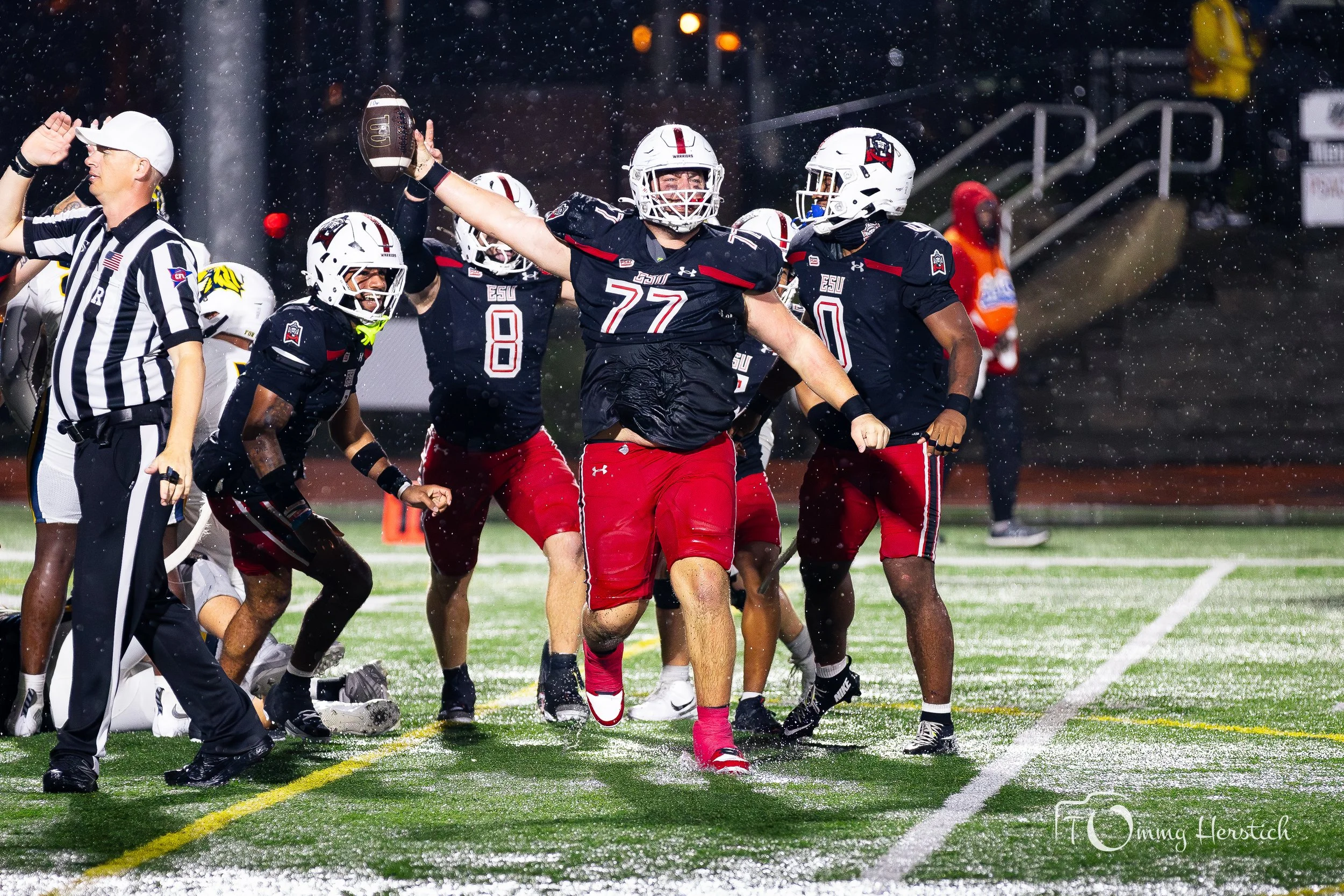 American football players celebrating on the field after a play, with a referee nearby, during a rainy night game.