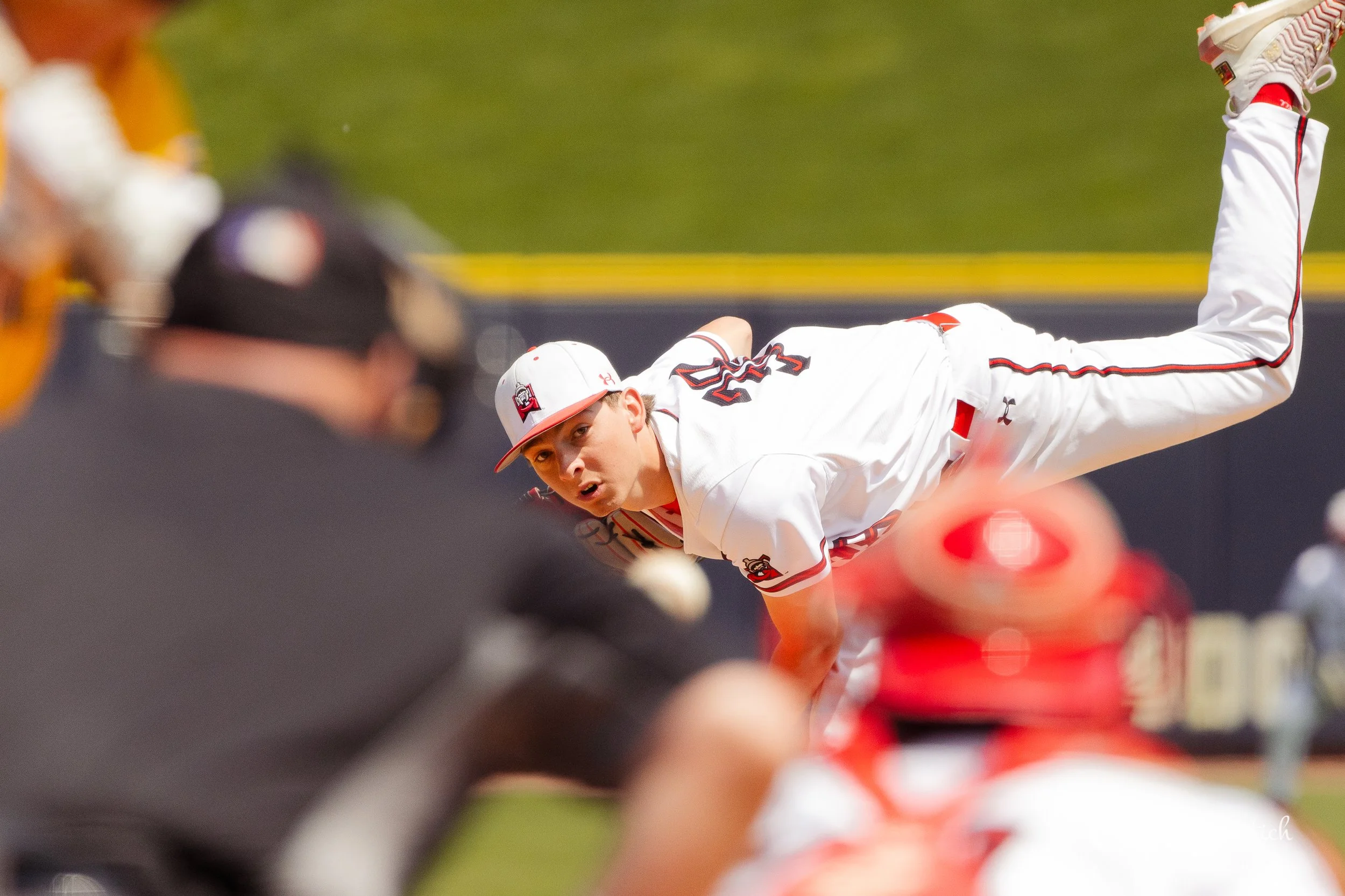 A baseball player in a white uniform and cap is diving on the field during a game, with an intense look on his face. The background shows a blurred outfield and a coach or umpire in black in the foreground, partially out of focus.