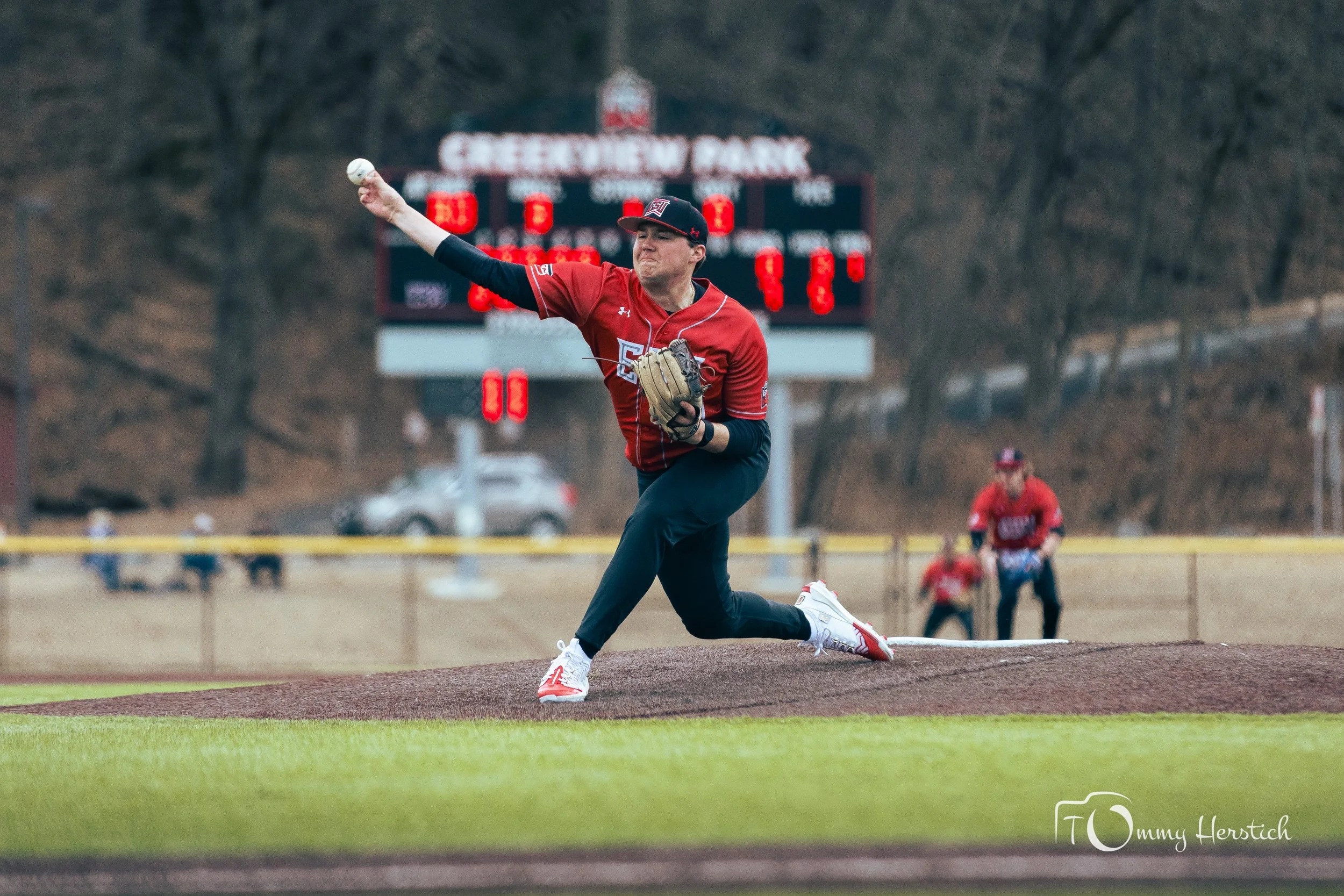 A baseball player in a red uniform and black pants is pitching the ball on a baseball field. The background shows a scoreboard and some players near the fence.