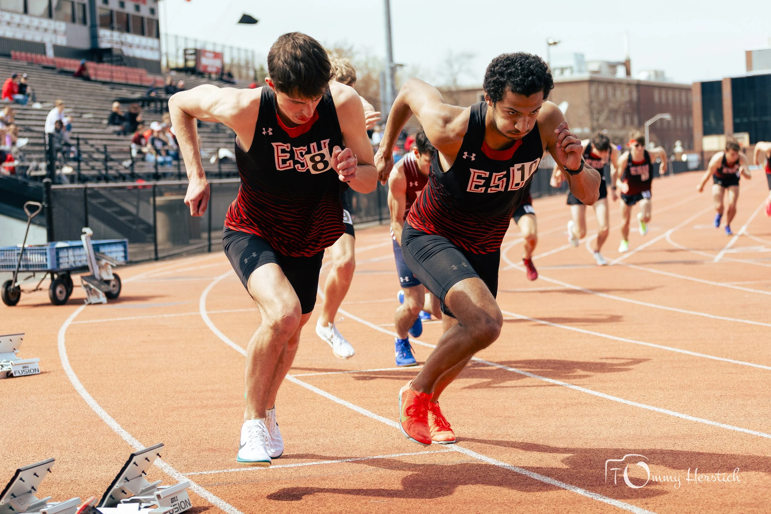 Two male athletes competing in a track race, preparing to start, on an outdoor stadium track with other runners in the background.