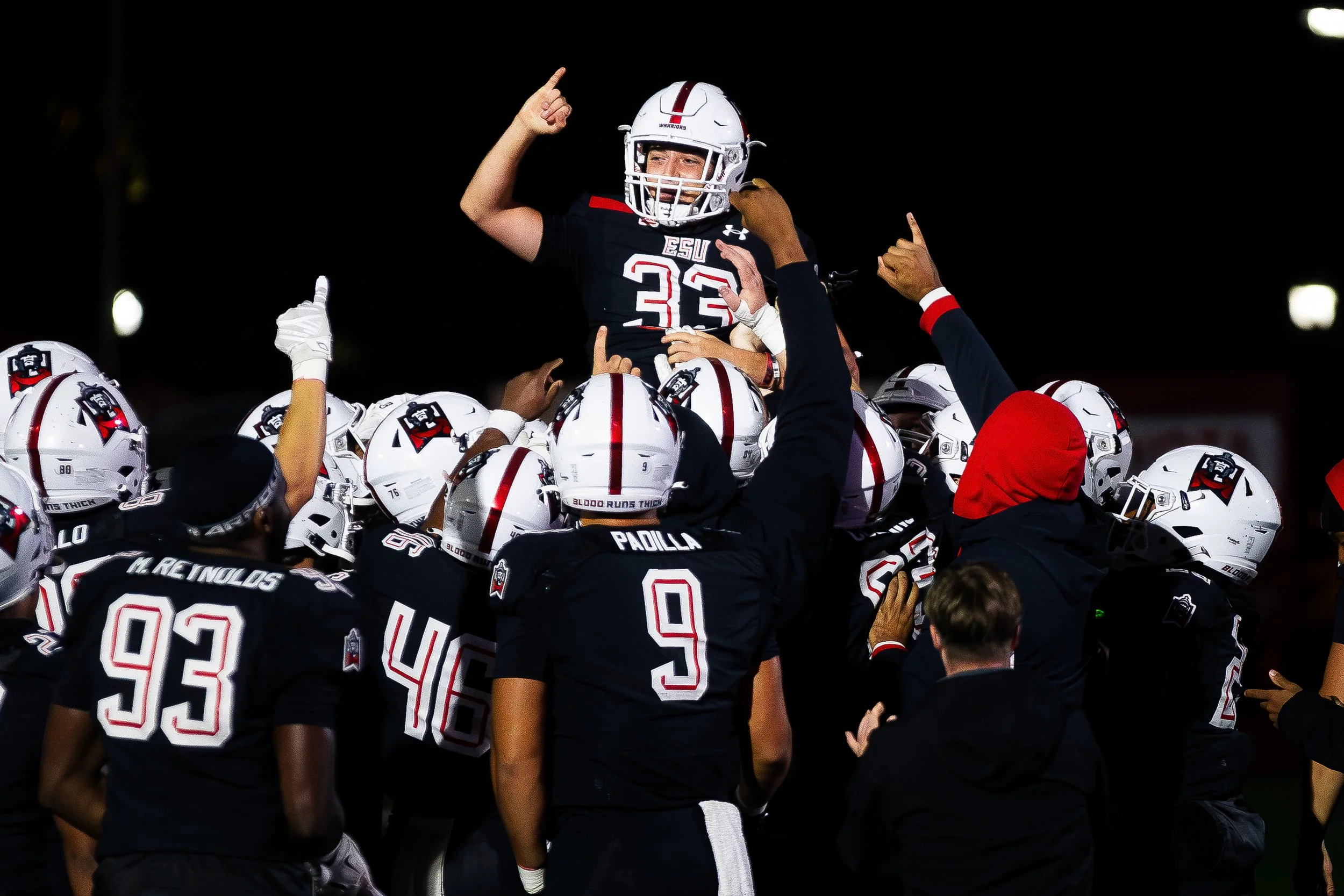 Football team celebrating at night, with one player being lifted by others, wearing black jerseys with red and white accents.