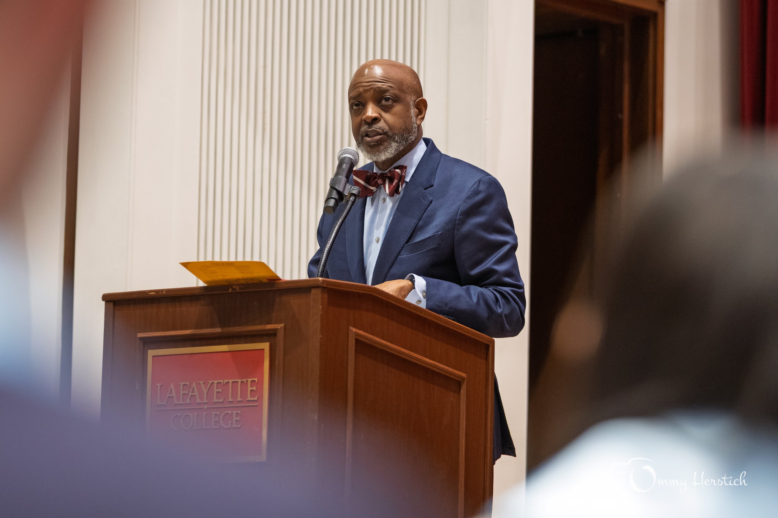 A man in a navy suit with a red bow tie speaking at a podium with a Lafayette College emblem.