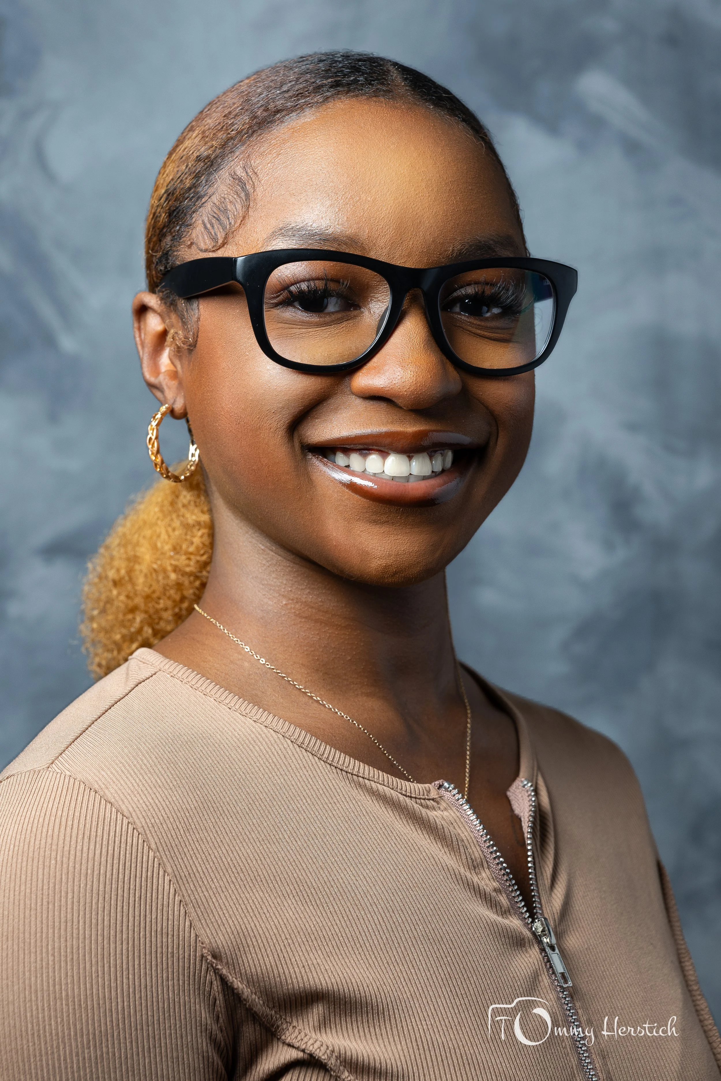A smiling woman wearing black glasses, hoop earrings, a beige zip-up top, and a delicate necklace, against a gray textured background.